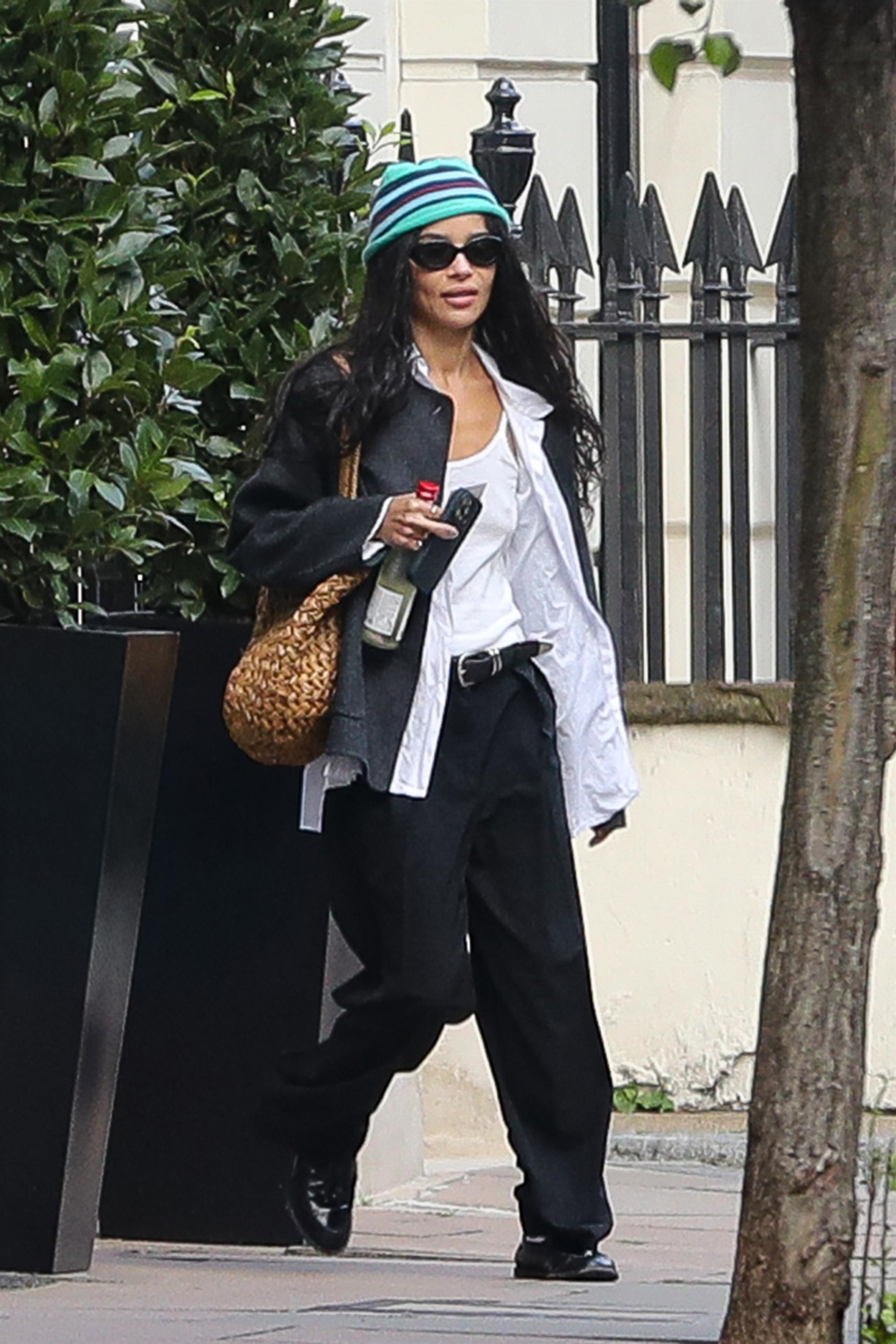 Zoe kravitz wears a stripe hat, black jacket, white shirt, tan raffia bag, black trousers, and black loafers.