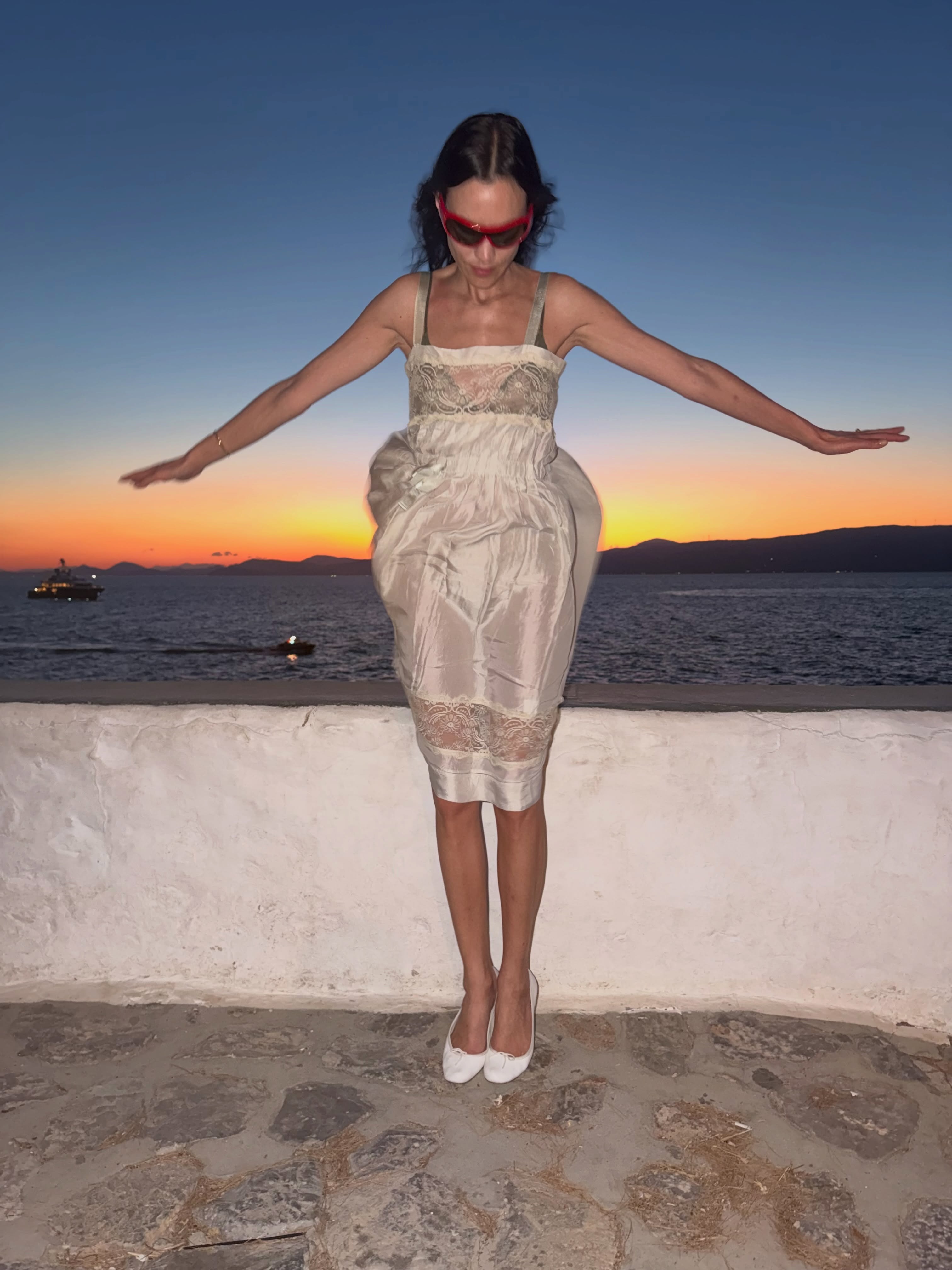 Alexa Chung wears a white lace-trim knee-length dress with white ballet flats and red sunglasses.