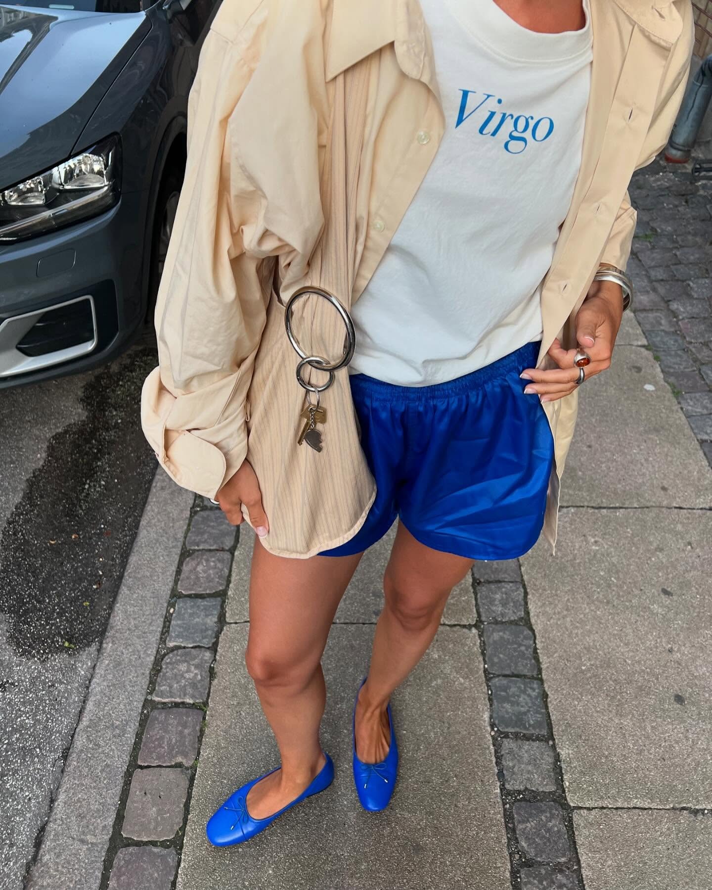 Danish style influencer Amalie Nielsen poses on a Copenhagen sidewalk wearing a chic 2026 outfit with a beige jacket, Virgo graphic T-shirt, bright blue shorts,  and royal blue ballet flats