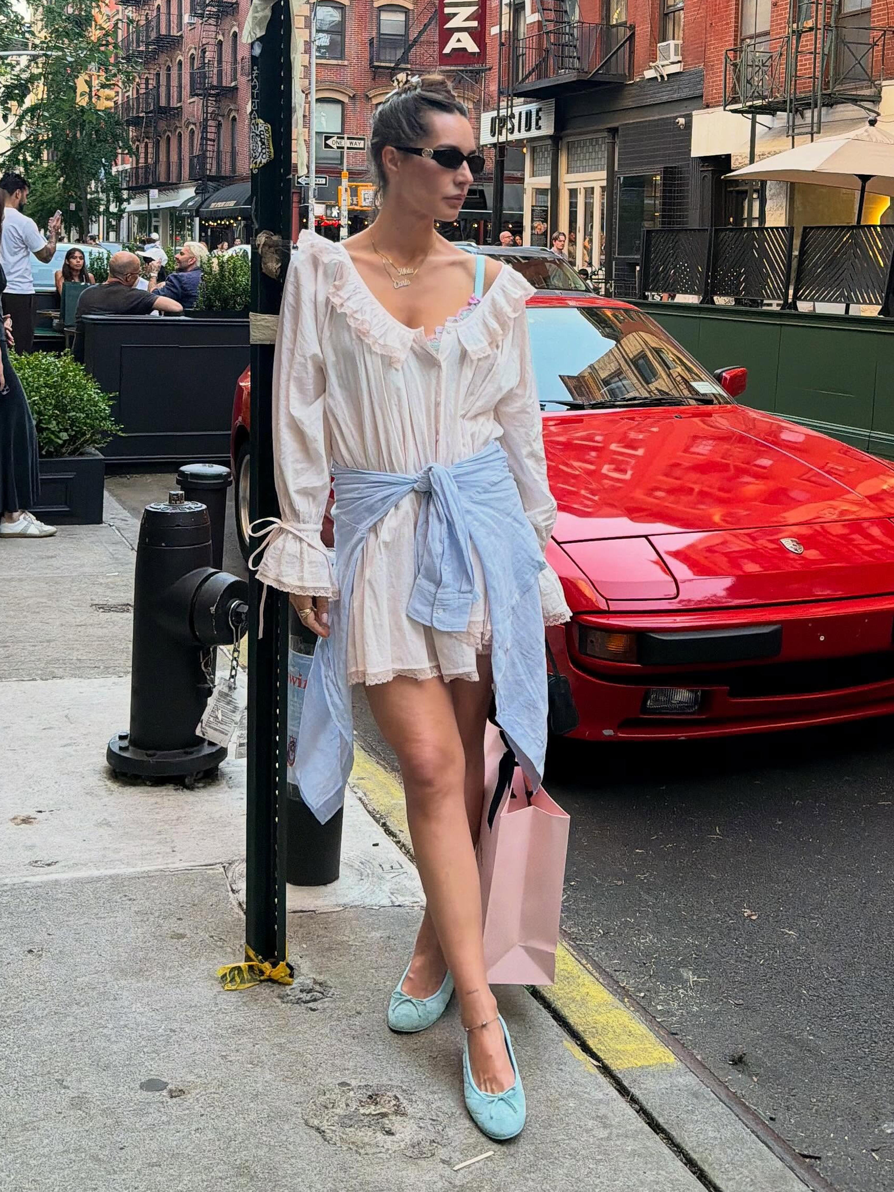 style influencer and designer Pia Baroncini poses on a sidewalk in NYC wearing a chic 2026 outfit idea with a ruffled minidress, blue linen shirt tied around the waist and blue ballet flats