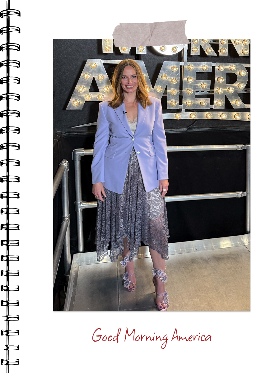 Carley Fortune poses in front of the Good Morning America sign wearing a purple blazer buttoned over a silver lace dress.