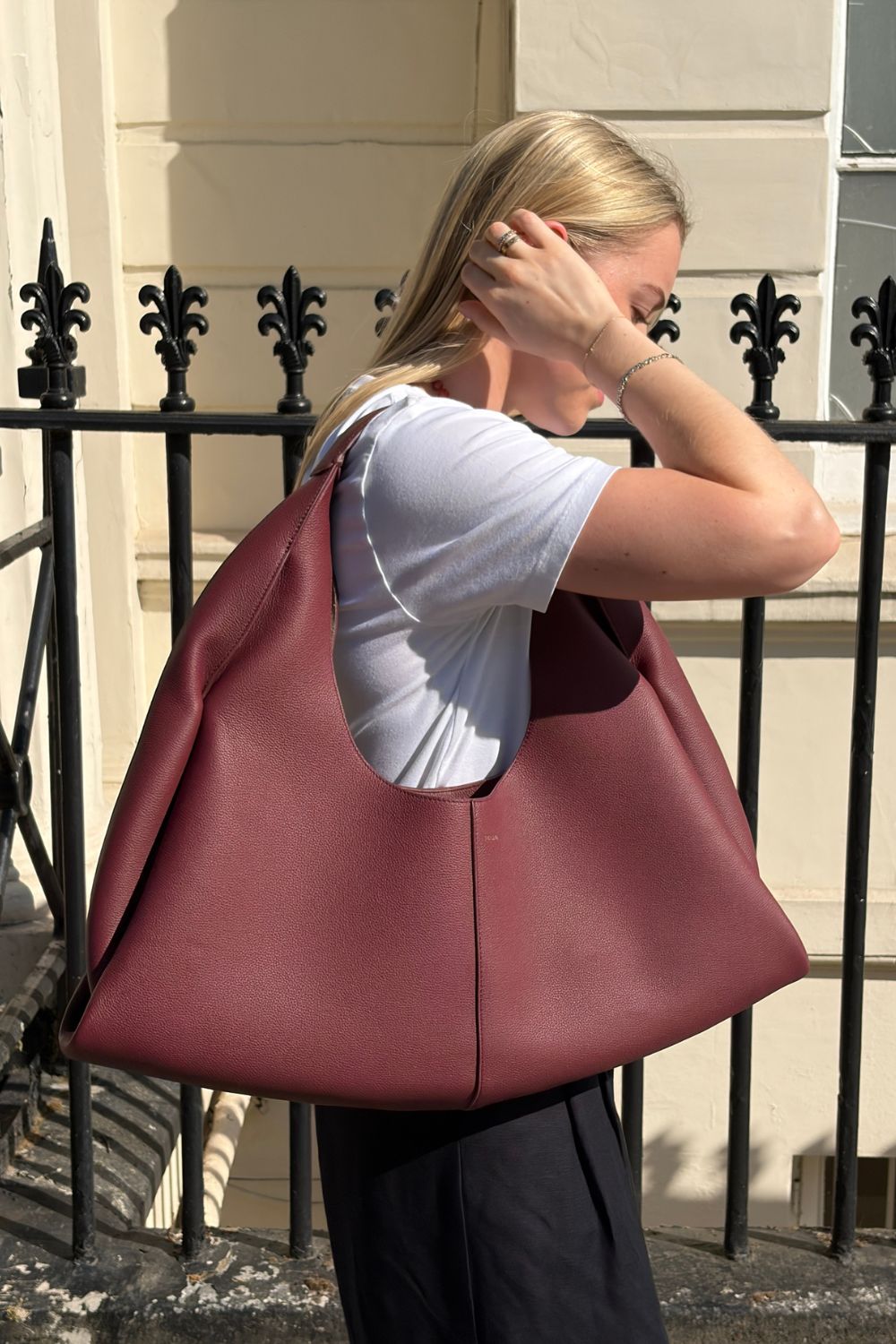 Woman wears white T-shirt, black linen trousers and carries burgundy Polene Shuldo bag