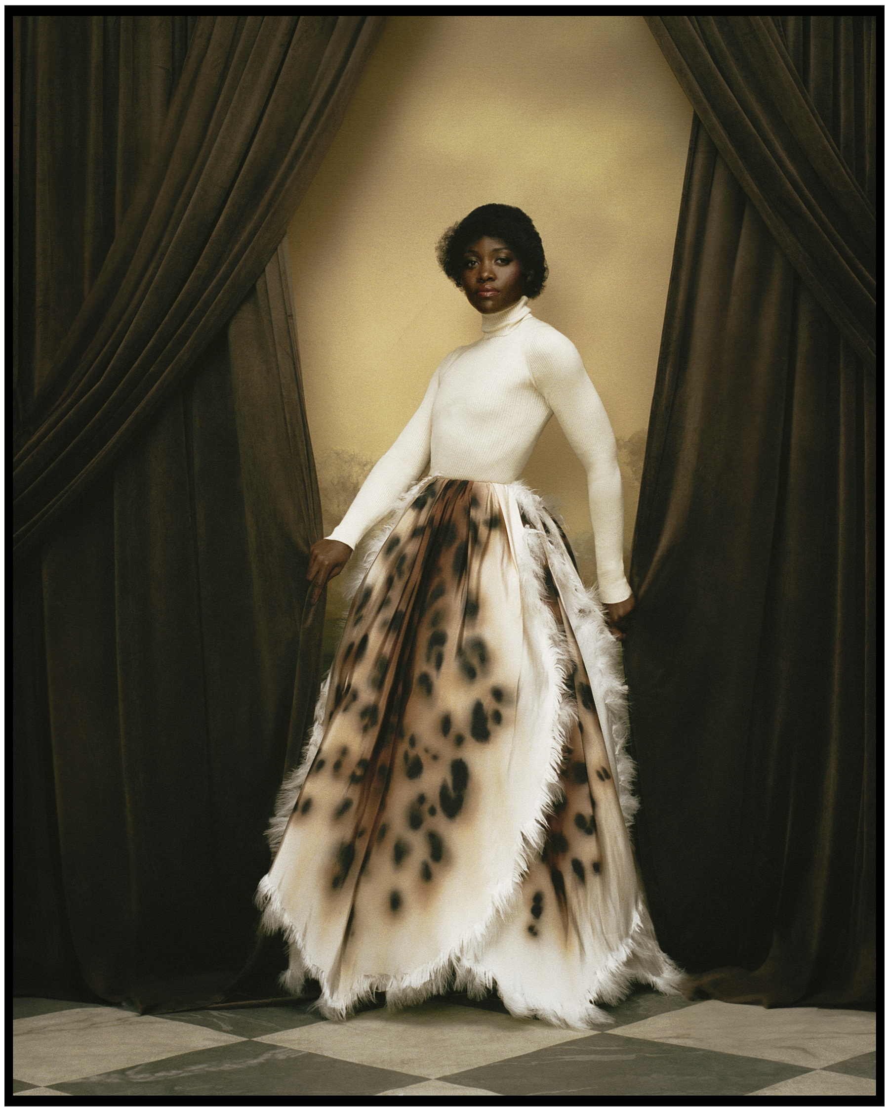 Lupita Nyong'o photographed by Kenny Germé standing between brown velvet curtains wearing a Chanel white turtleneck tucked into a full skirt with leopard print design.