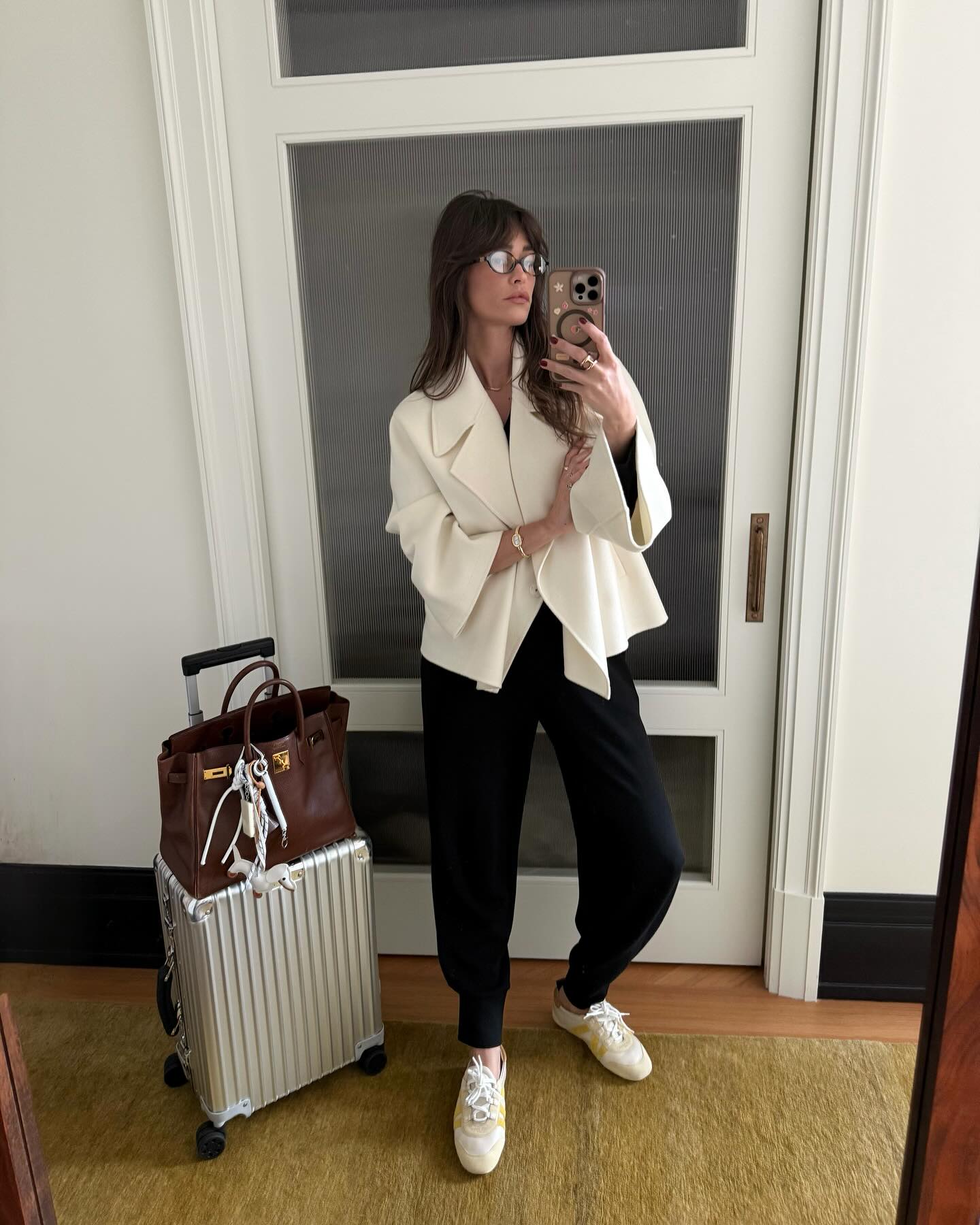 An image of @alisontoby wearing a rich-looking airport outfit including a white shawl, black joggers and low-rise trainers.