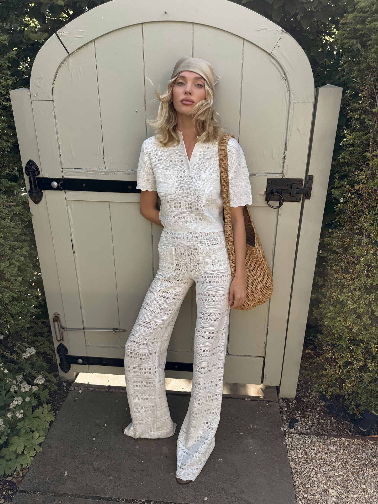 An image of a model wearing a white crochet T-shirt, matching crochet trousers and a beige hair bandana standing in front of a gate wearing a woven bag