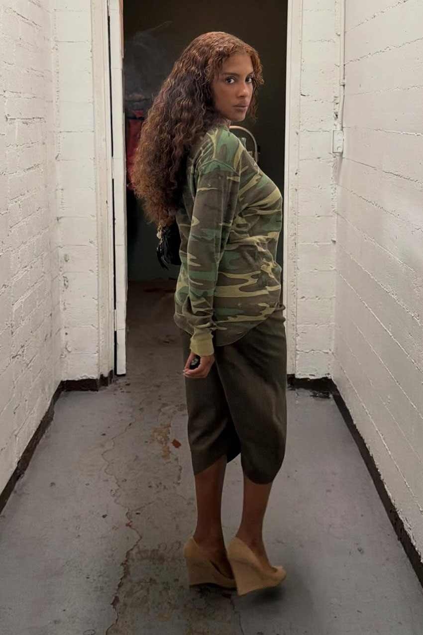 A woman wearing a camo long-sleeve, green shorts, and nude wedges.