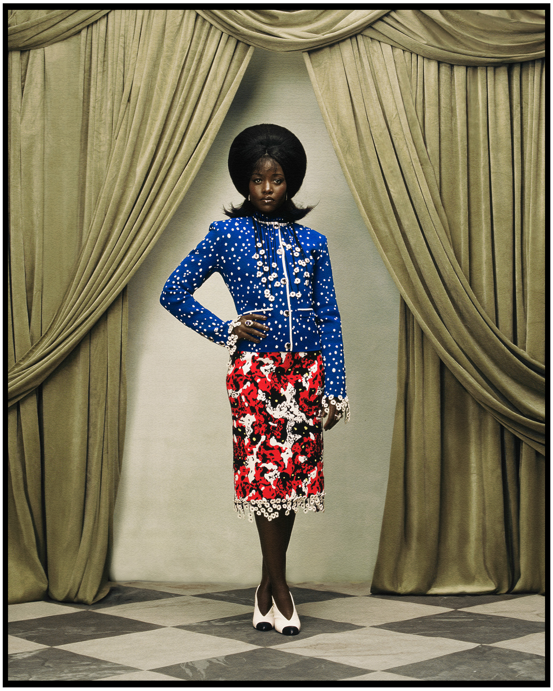 Lupita Nyong'o photographed by Kenny Germé. She is standing in front of a velvet curtain wearing a Chanel blue jacket with white dots and a red pencil skirt.