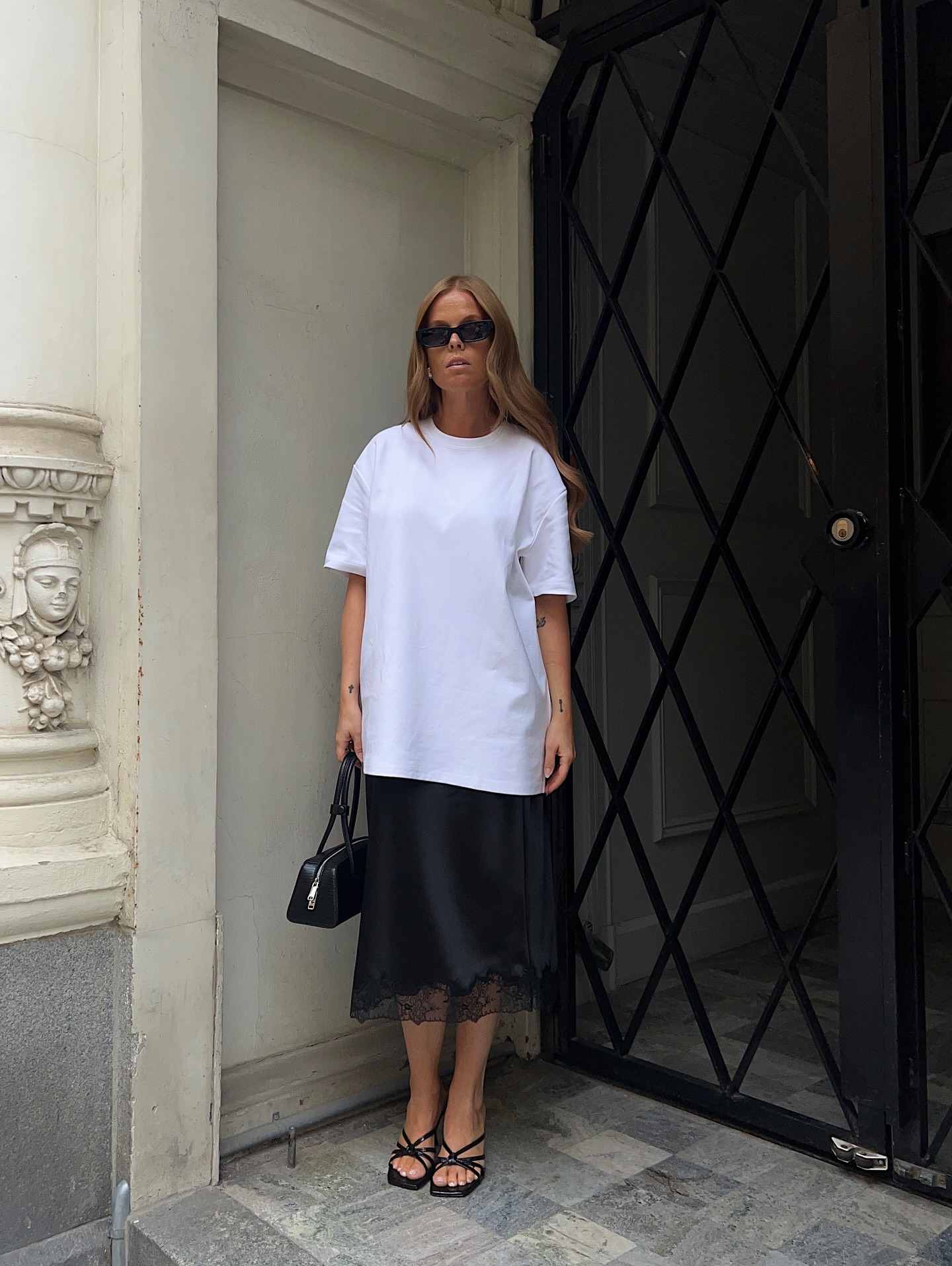An image of an influencer wearing a long white T-shirt over a black, lace-trim slip skirt and heeled black sandals in Copenhagen