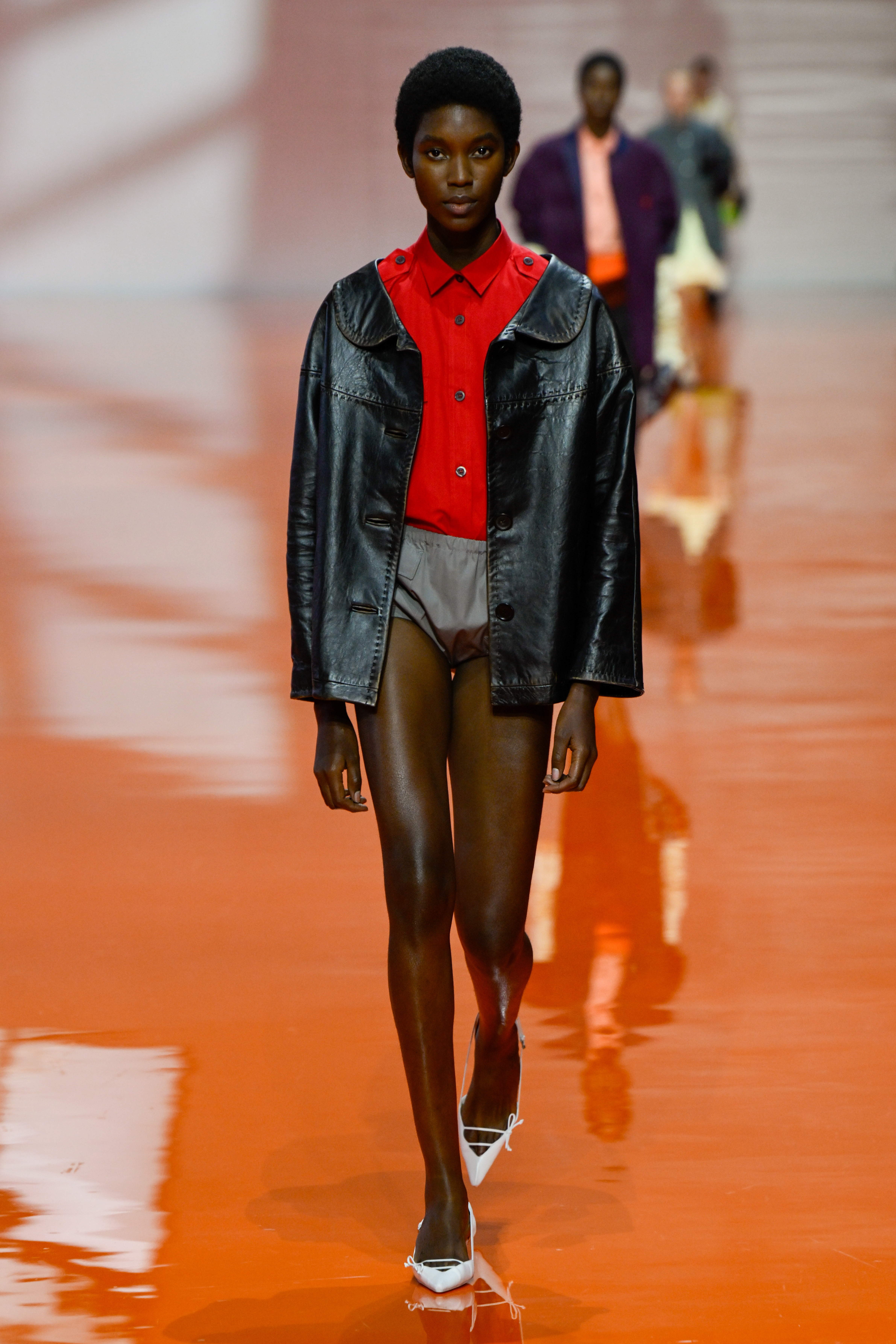 A model wears a red shirt and a black leather jacket on the Prada SS26 runway in Milan.