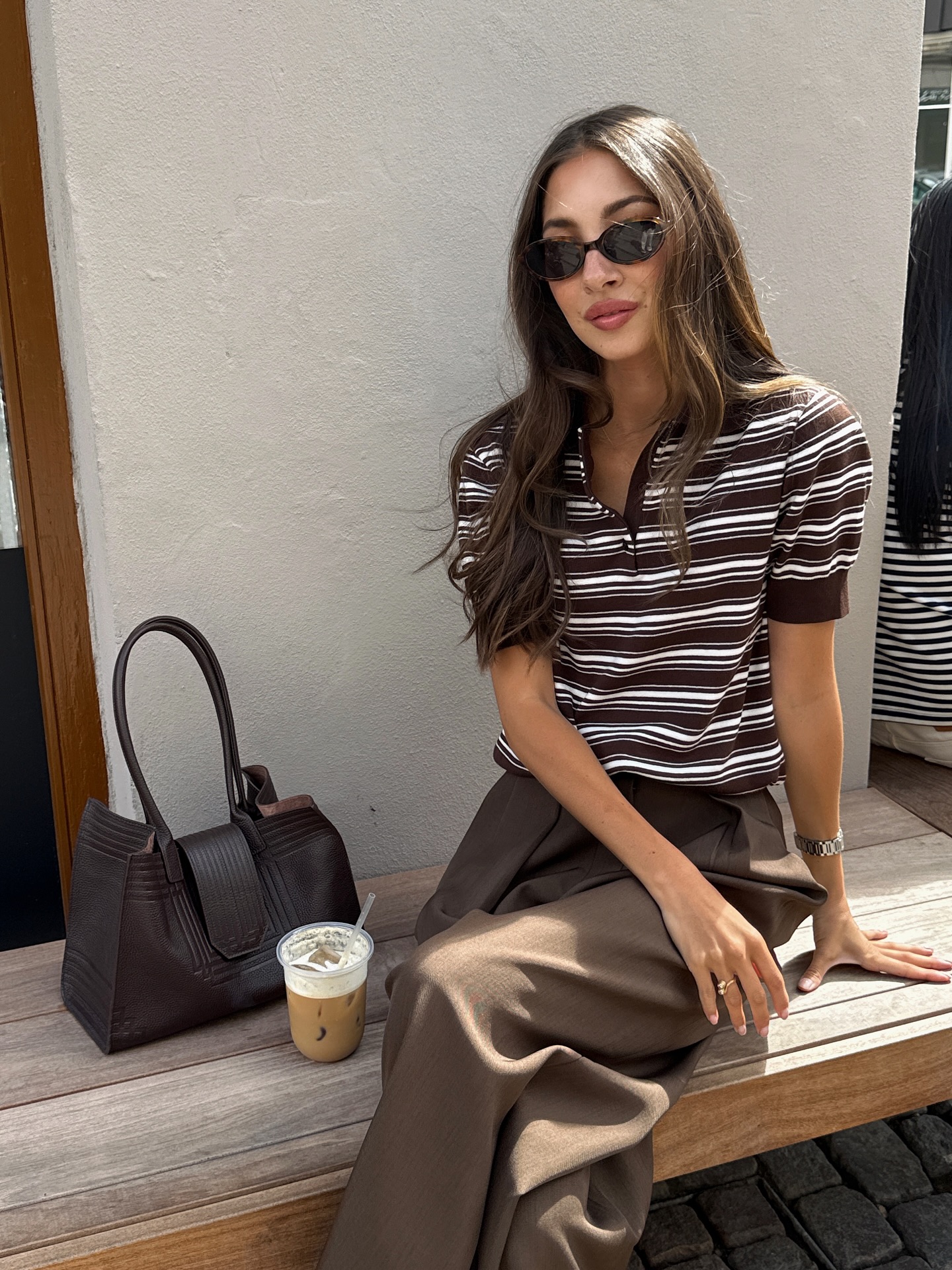An image of an influencer sitting on a bench in brown trousers and a brown and white striped T-shirt with a structured handbag in Copenhagen