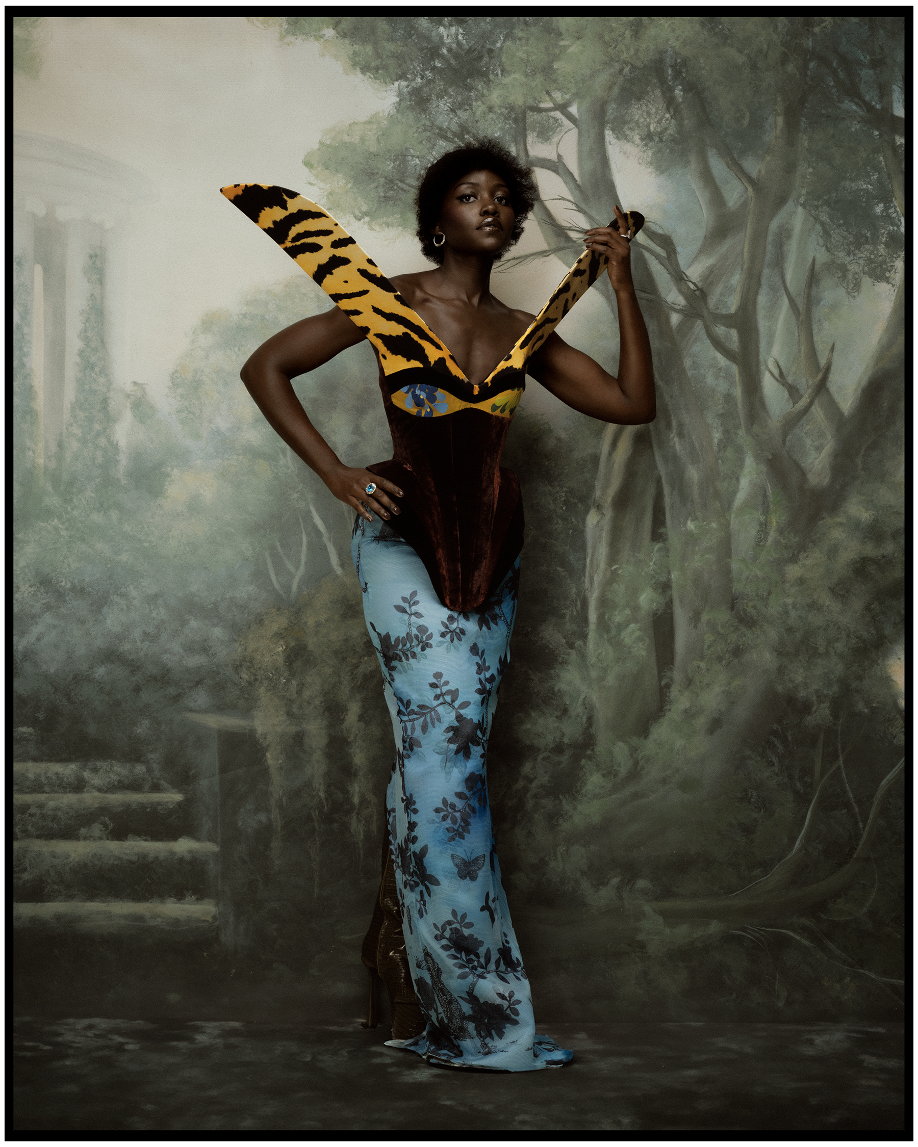Lupita Nyong'o photographed by Kenny Germé wearing a gown by Harris Reed. She is standing in front of a backdrop with a painted forest scene.