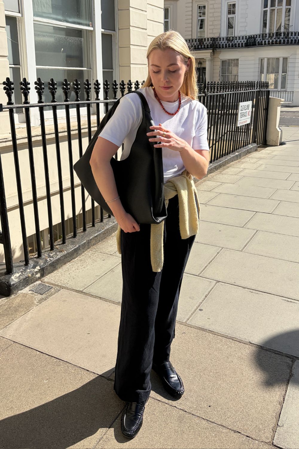Woman wears white T-shirt, red necklace, black linen trousers, carries black Shuldo bag by Polene