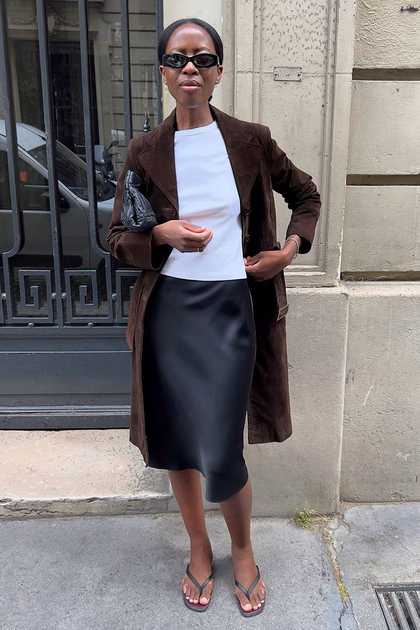 a woman wearing a white tee, black satin skirt, brown coat, and flip-flops