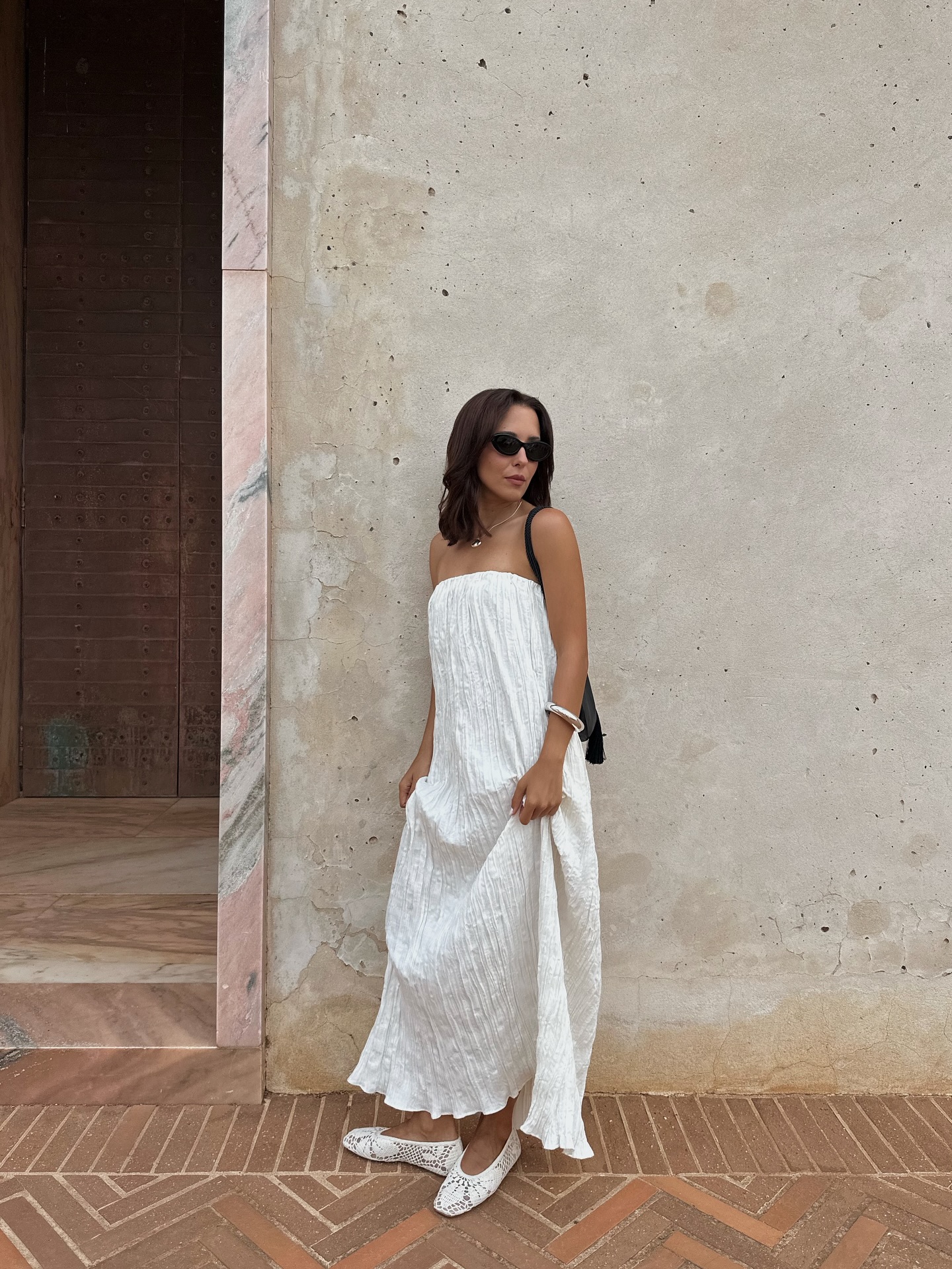 An image of an influencer wearing a white strapless dress and white crochet flats in front of a concrete building