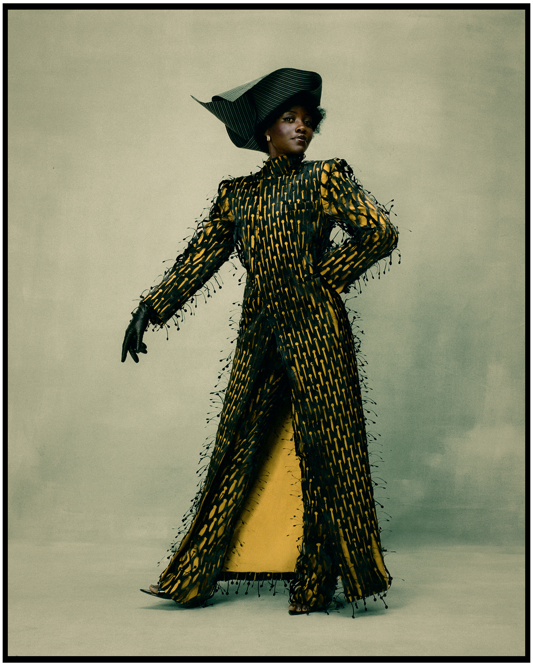 Lupita Nyong'o photographed by Kenny Germé standing in front of a painted gray/blue backdrop wearing a black and yellow floorlength duster coat with matching pants and a black structured hat.