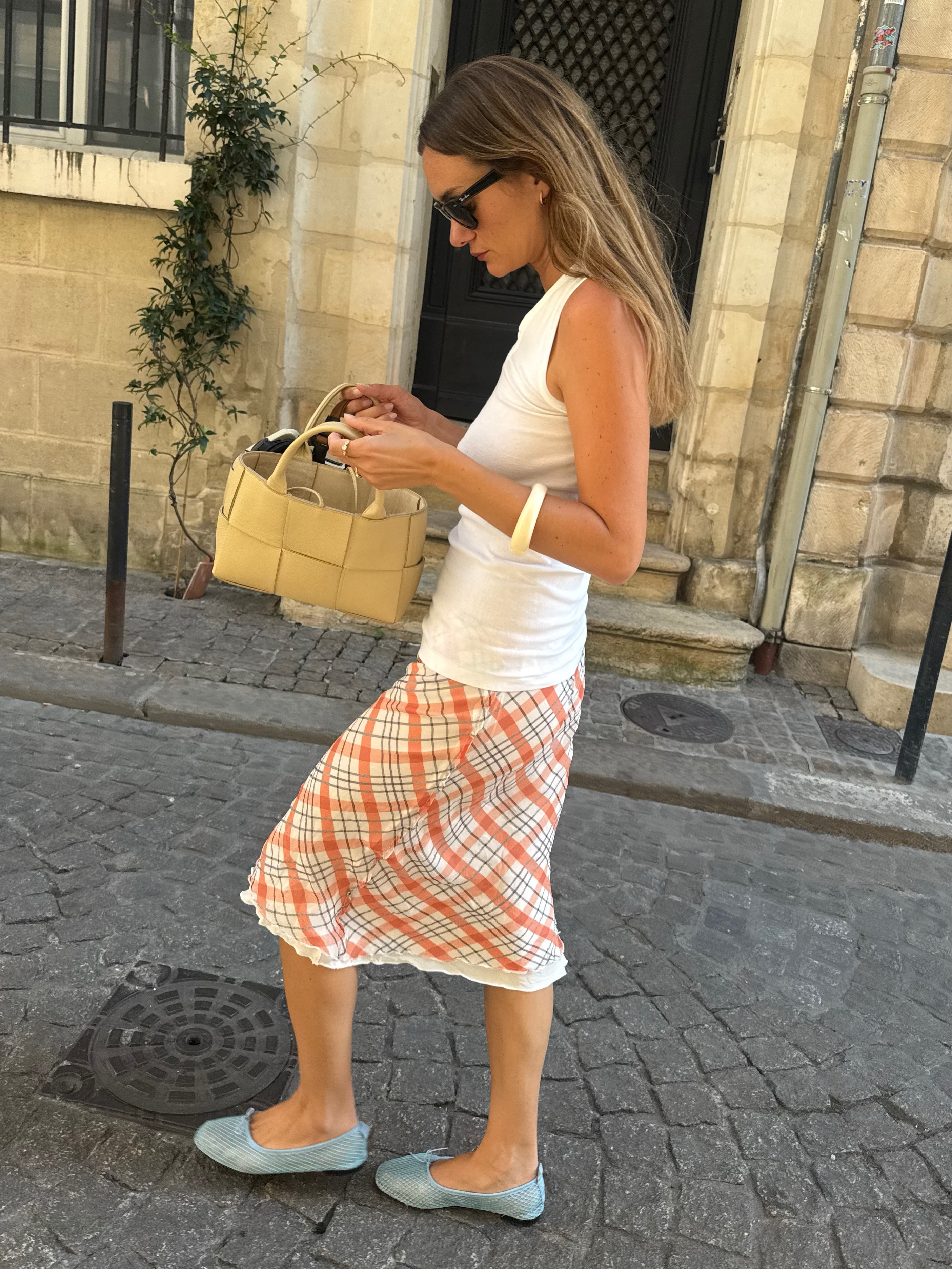 An image of an influencer wearing a white vest, plaid skirt and aqua mesh ballet flats in Paris