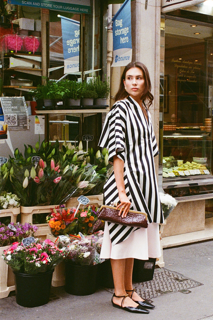 A woman wearing a long black-and-white striped tunic shirt styled over a white shirt, black pointed ballet flats, and a red clutch.