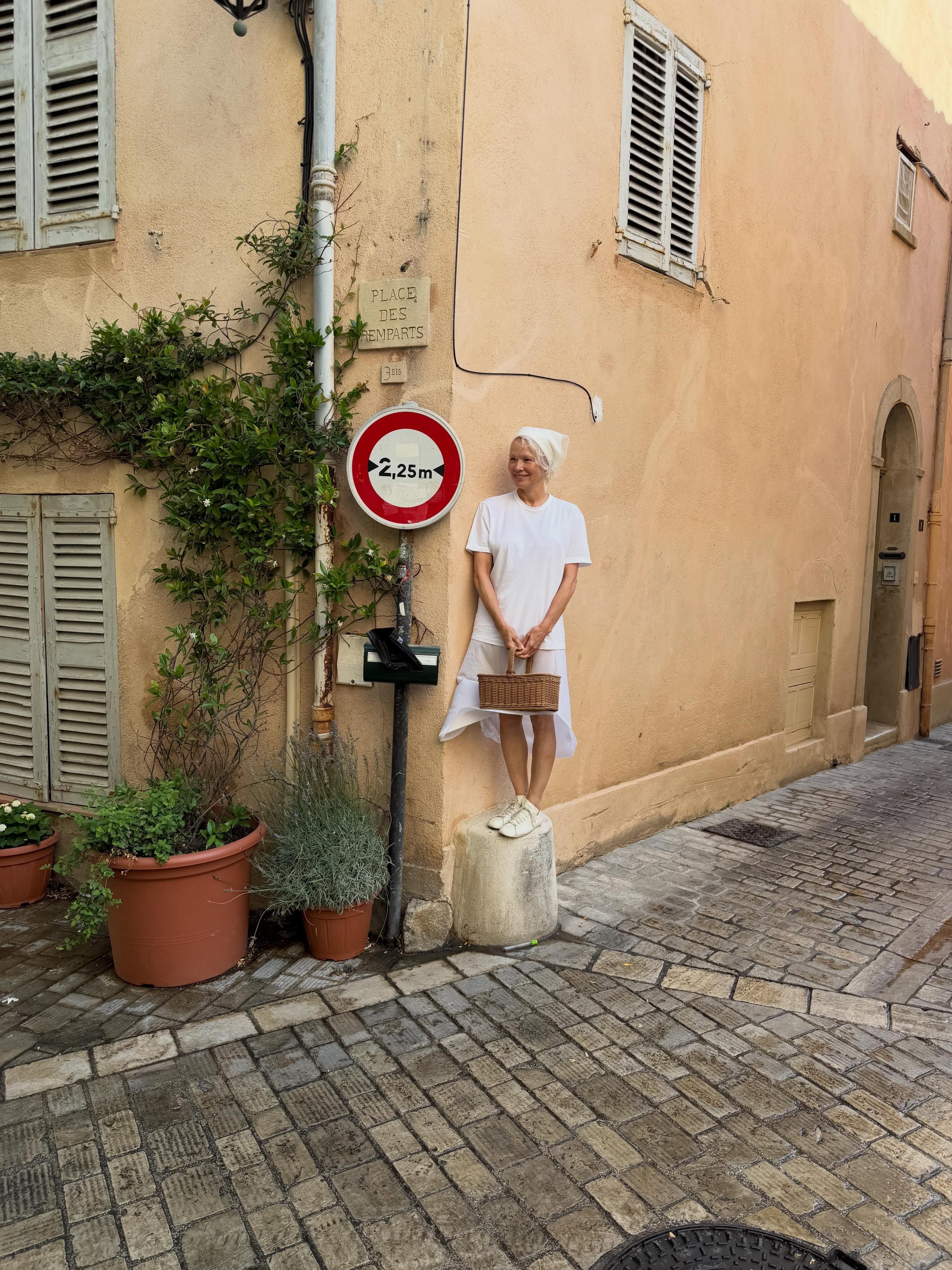 Pamela Anderson wears a white midi dress with a white headscarf, a raffia bag and white flats.