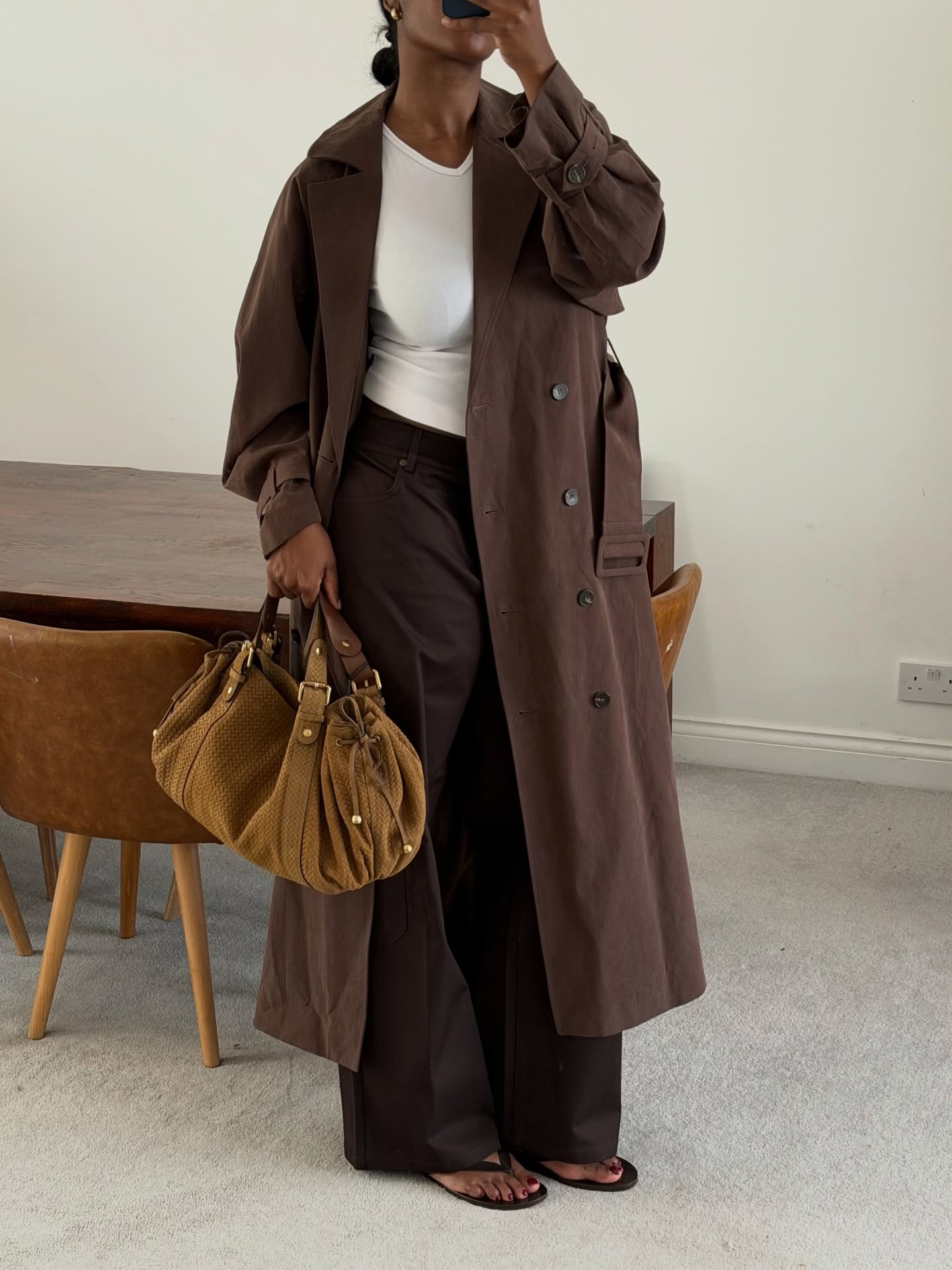 Woman wearing a brown trench coat, a white tee, and relaxed brown trousers.