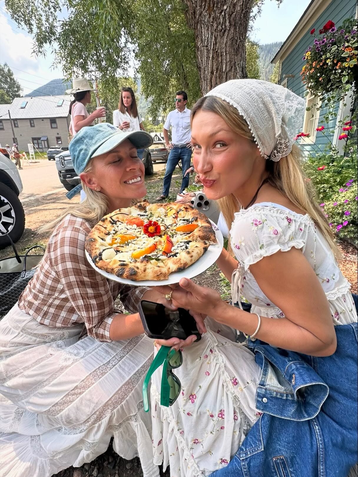 Bella Hadid holds up a pizza wearing a cotton floral dress with a white bandana.