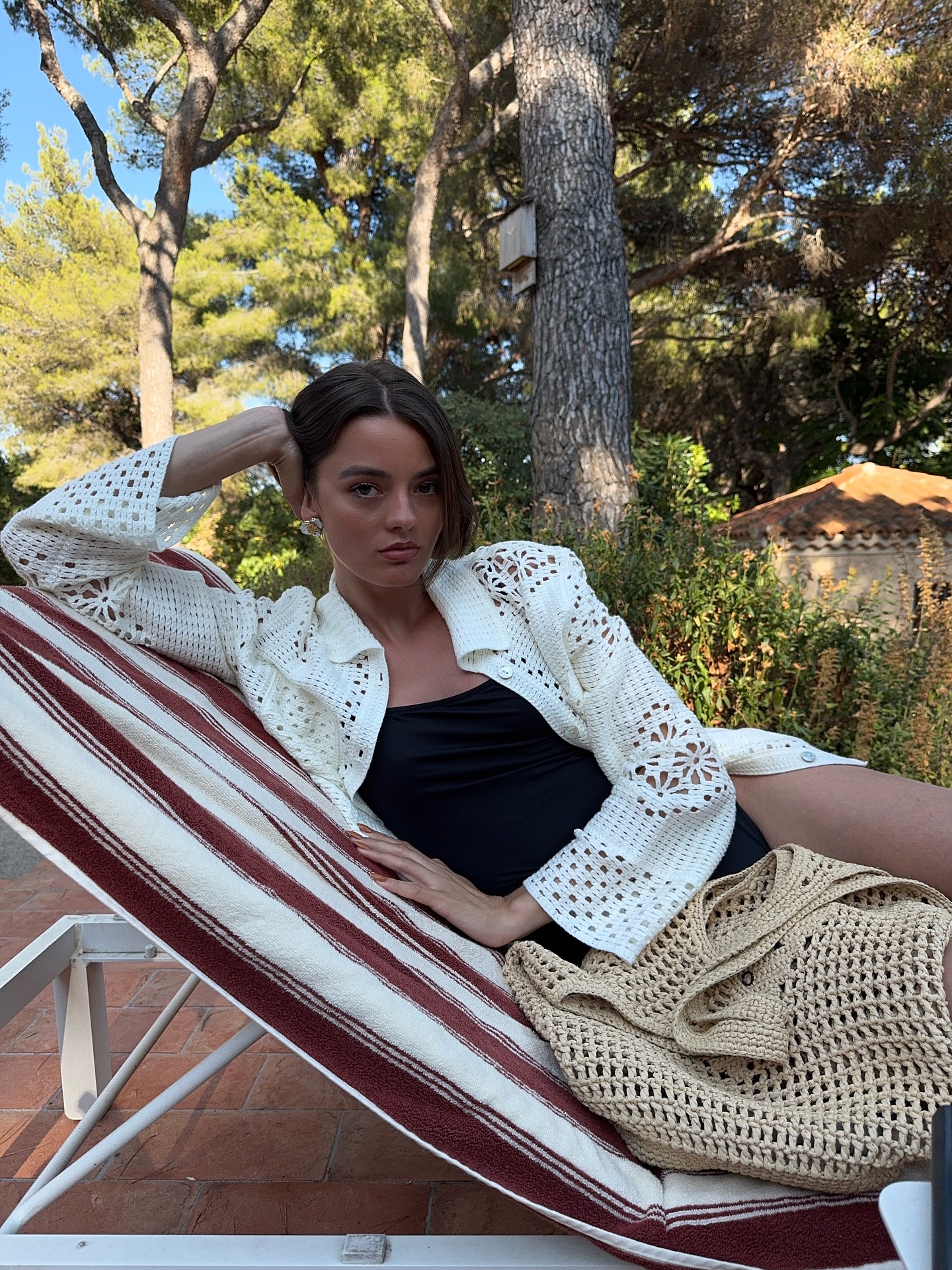 An image of an influencer wearing a white crochet long-sleeve top on a sun lounger and a beige crochet bag in front of her