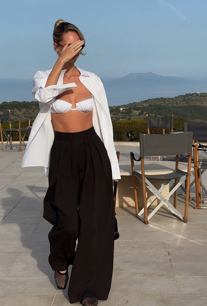 classic swim trend shown on a French woman standing on a patio near the sea wearing an open white button-down shirt with a white balconette bikini top, black white leg linen pants, and black flats