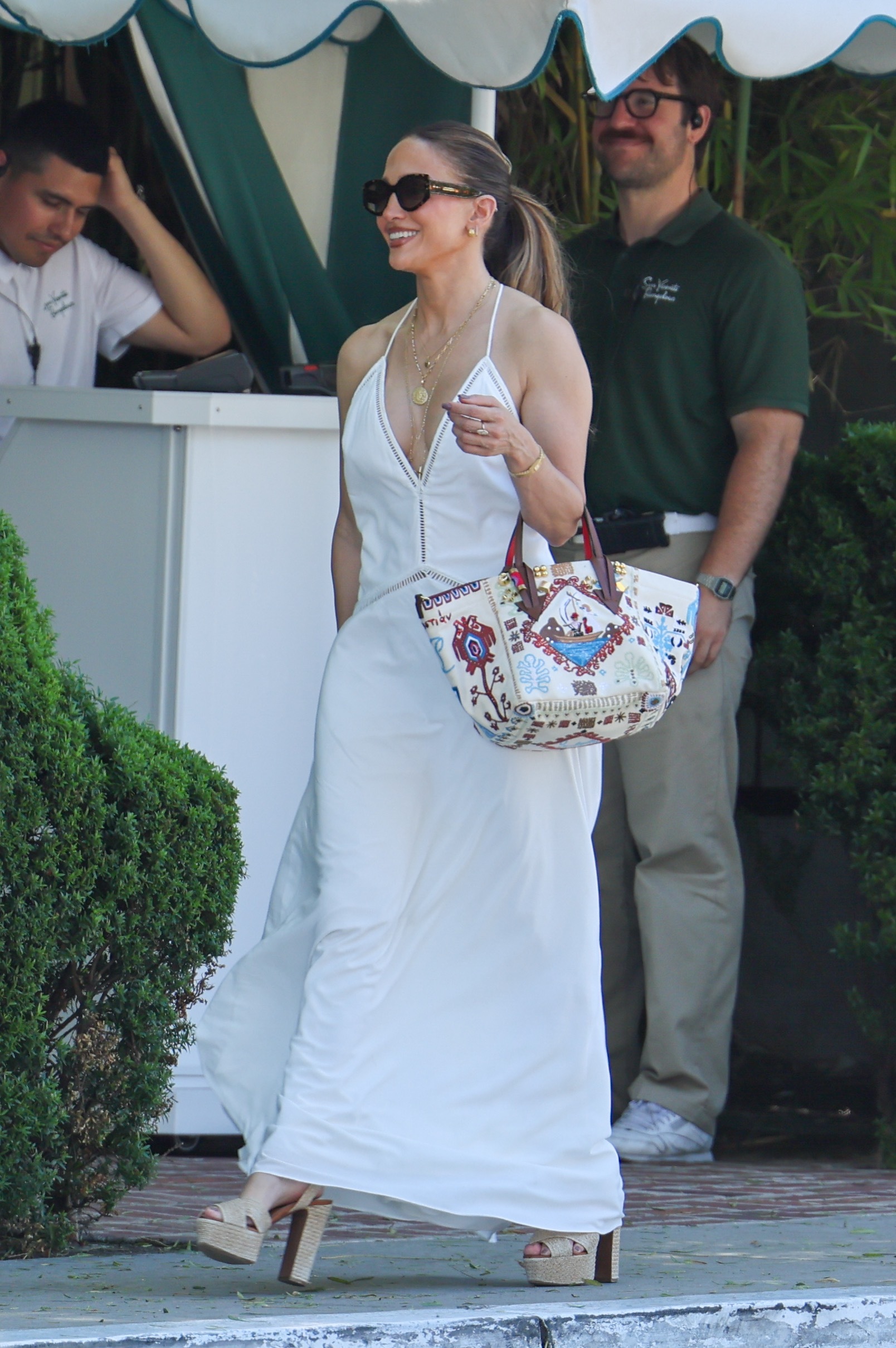 Jennifer lopez wears a white maxi dress, printed tote bag, layered necklaces, and raffia platform heels.