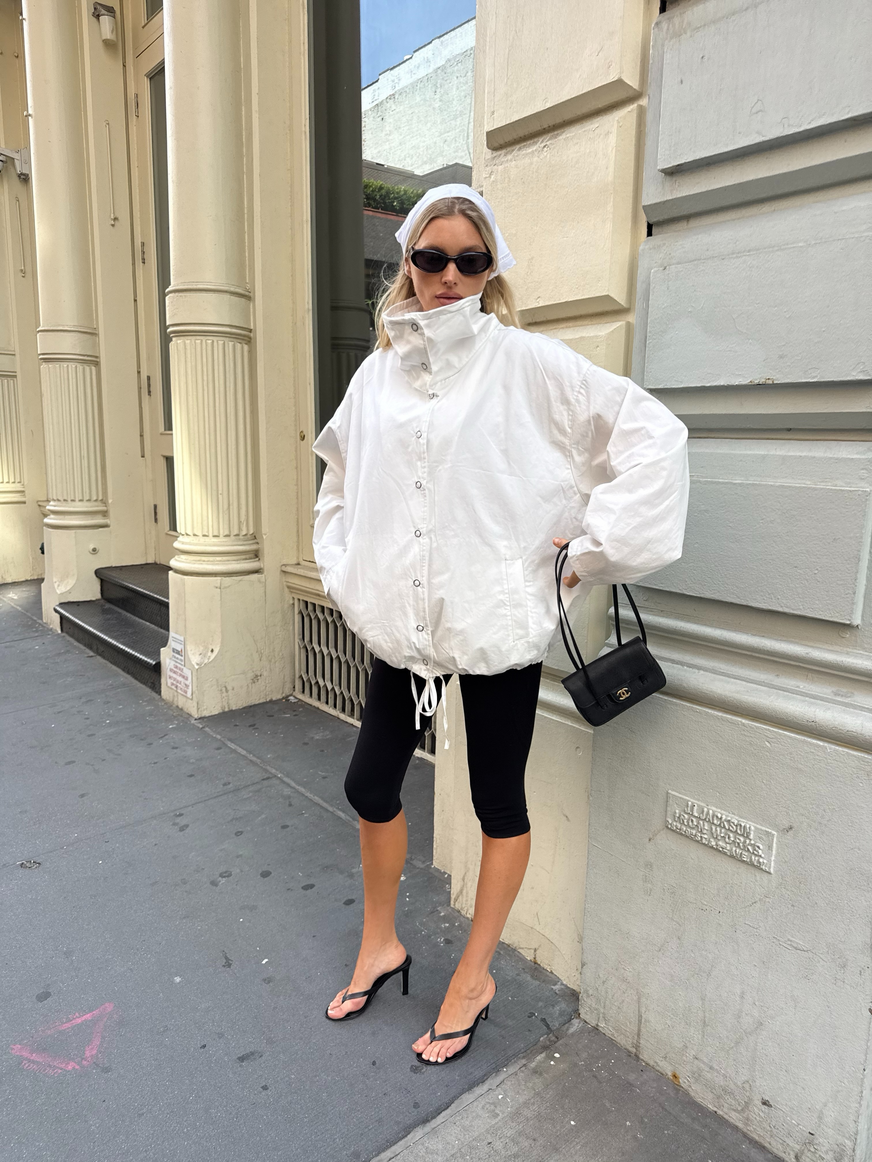 An image of an influencer wearing a white bandana in her hair, a white funnel-neck windbreaker, black capri leggings and black heeled flip-flops