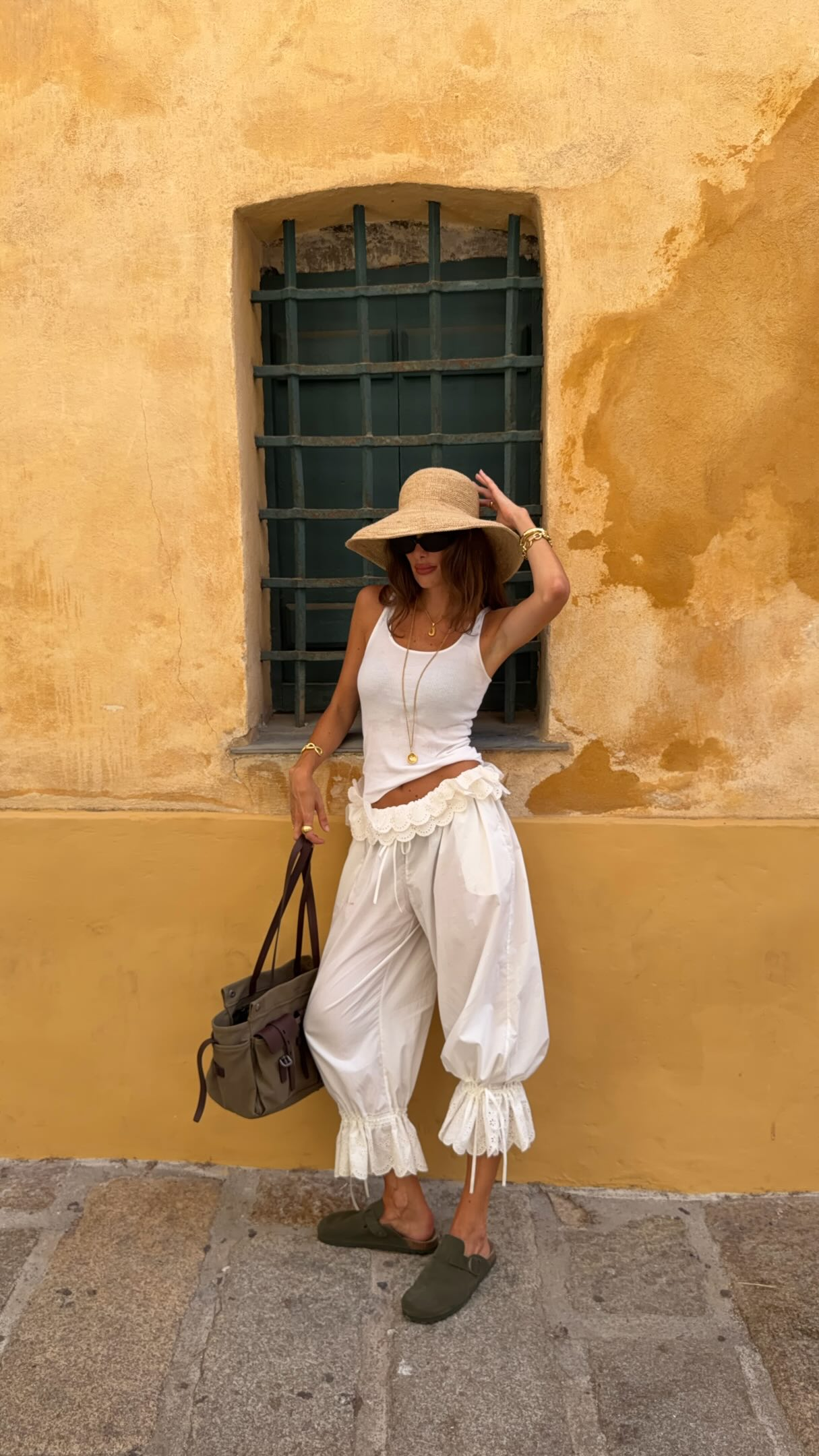 Influencer @juliesfi wears white bloomer trousers with a white tank top and a wide-brim straw hat.