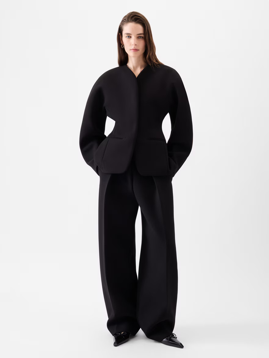 An image of a model wearing a rounded sculptural jacket in black with black trousers and pointed-toe heels