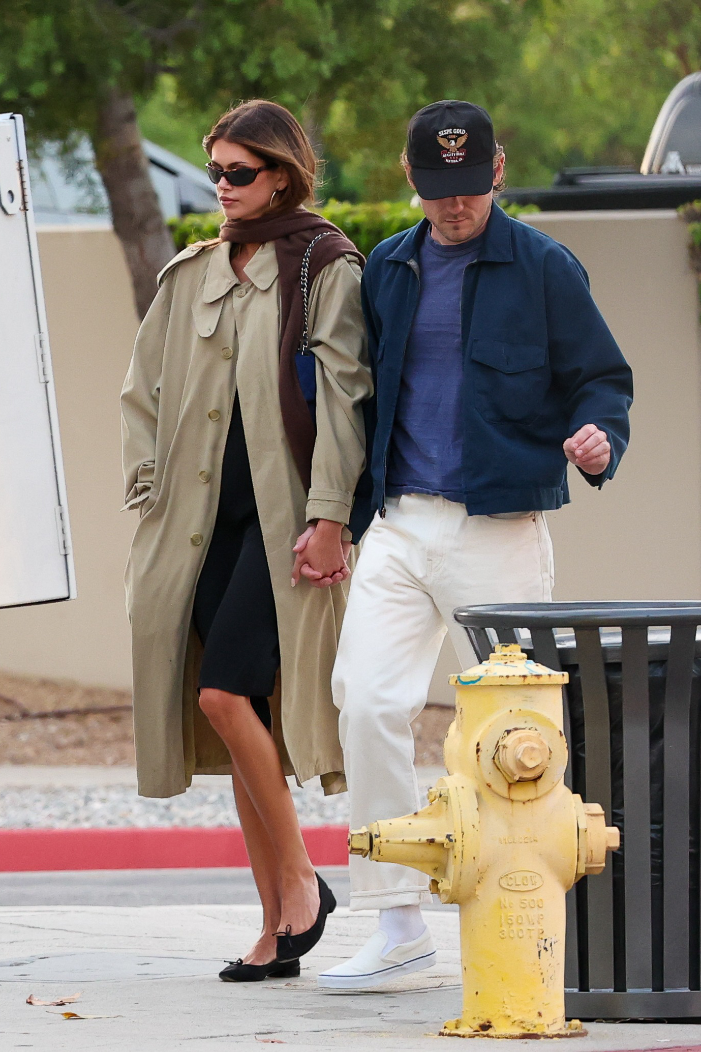 An image of Kaia Gerber wearing a trench coat with a black dress and ballet flats.