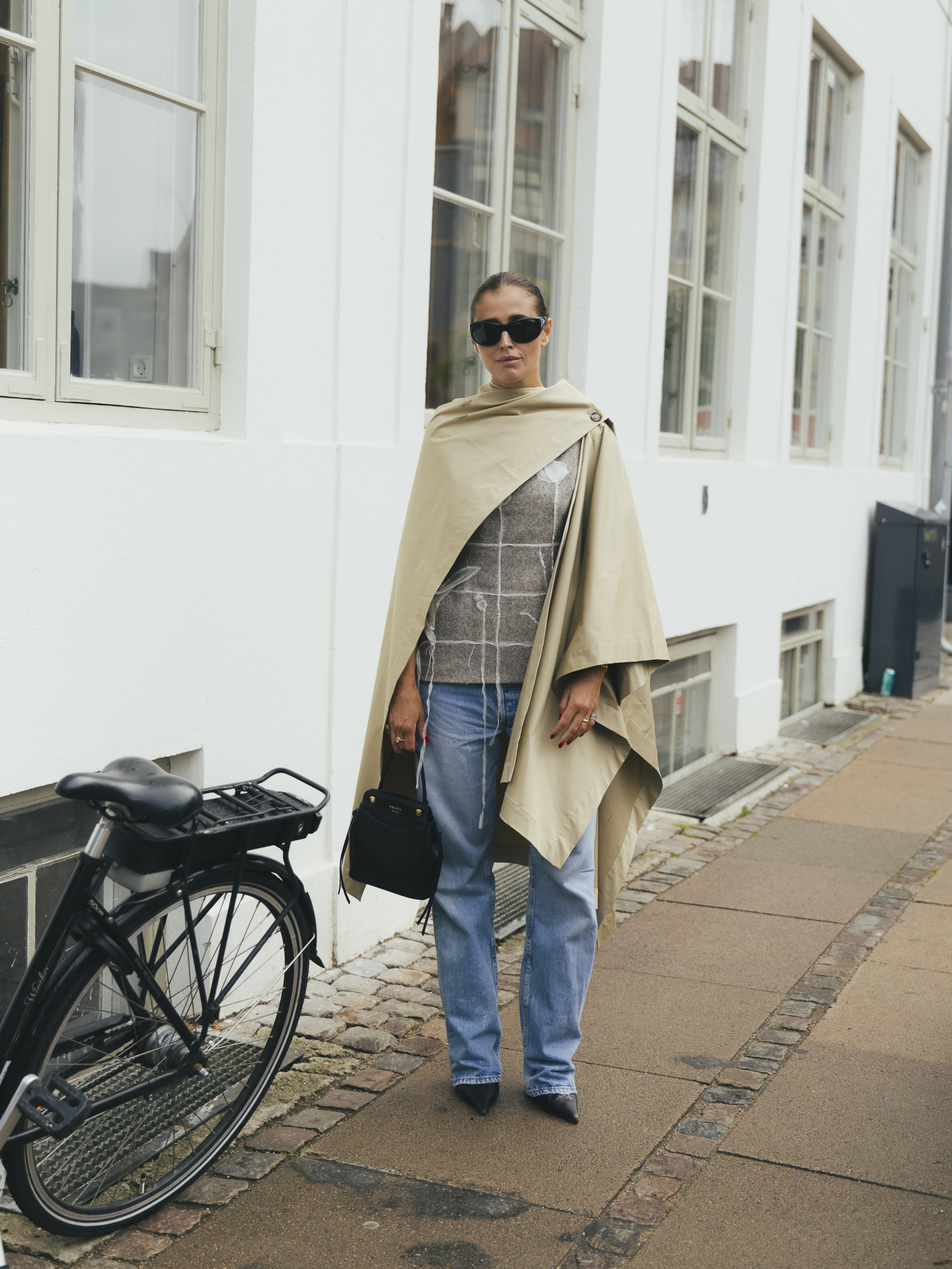 a woman wears a beige cape, straight-leg jeans, and black pointed toe heels.