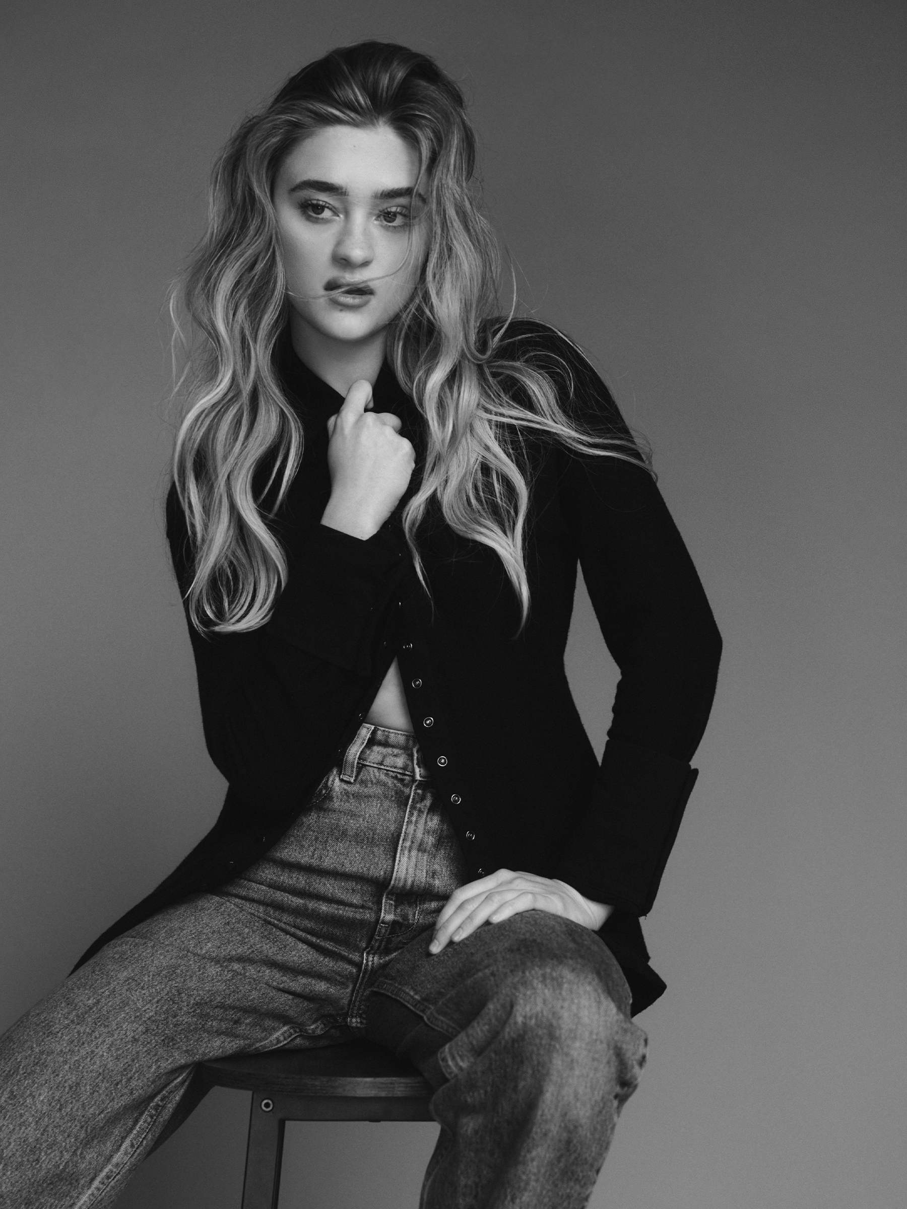 Lizzy Greene photographed by Raul Romo sitting on a metal stool wearing a long black cardigan with jeans.
