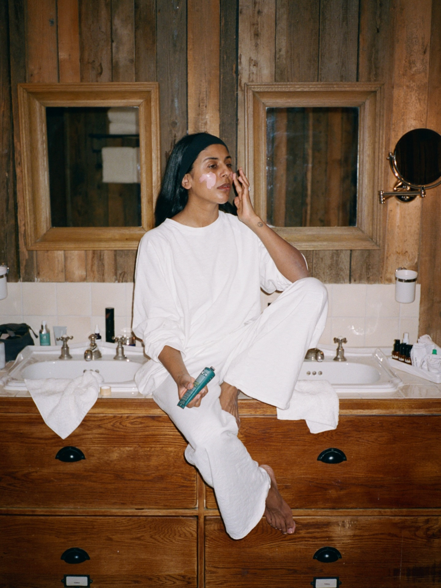 @monikh sitting on a sink surrounded by skincare products, applying a lotion to her face