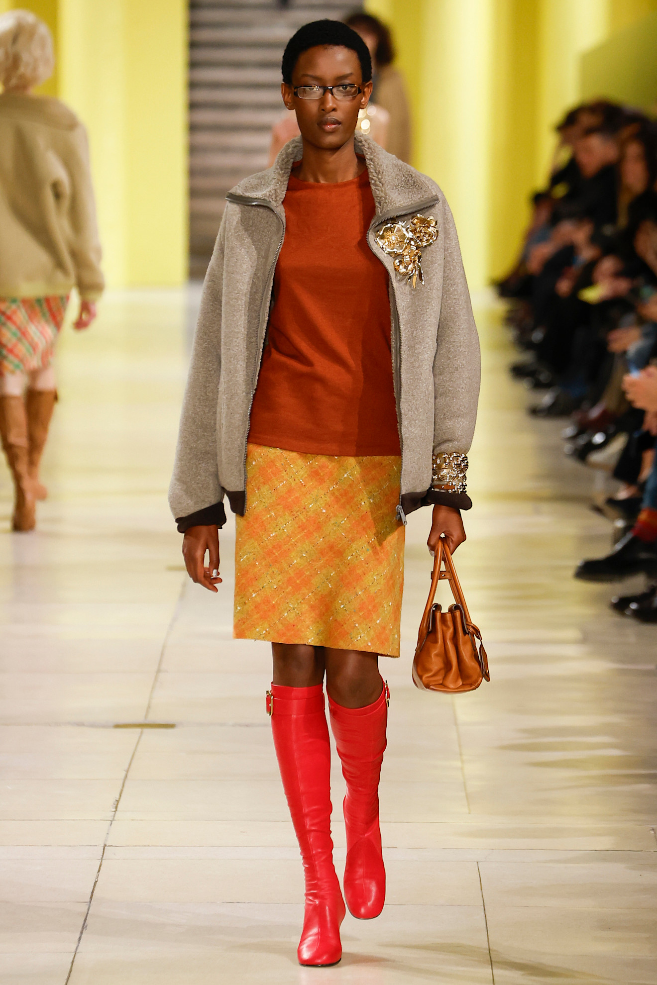 An image of a model wearing a beige coat, orange top, plaid orange skirt, and bright red boots on the Miu Miu autumn 2025 runway