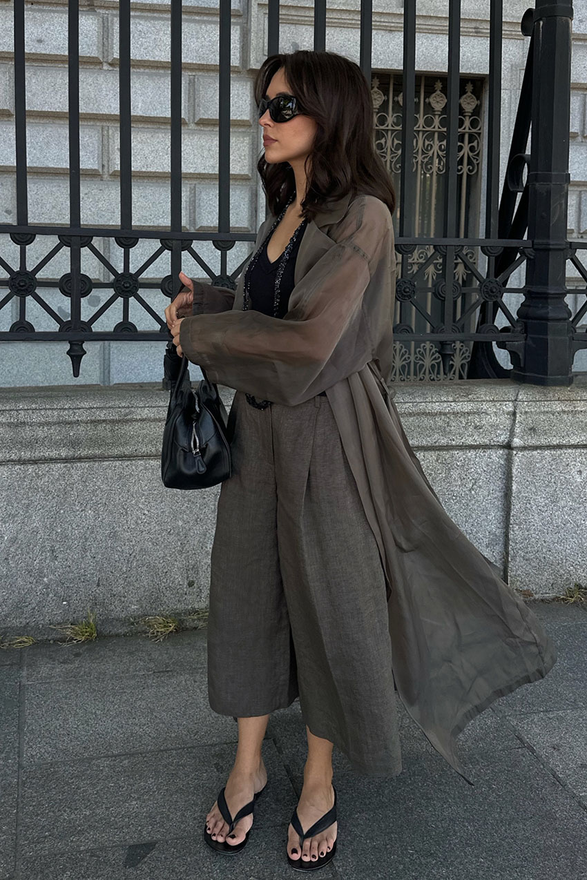 a woman wearing a gray trench coat, V-neck black tank top, gray long shorts, and thong sandals