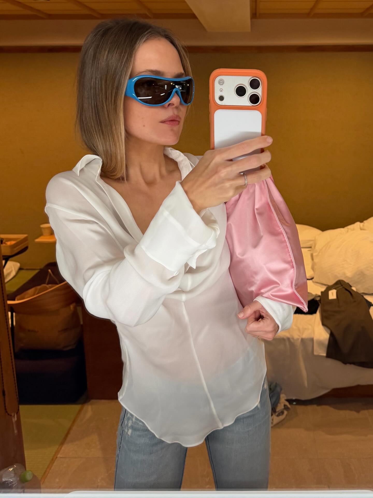 An image of an influencer wearing blue Miu Miu sunglasses, a sheer white blouse, blue jeans and a pink silk bag.