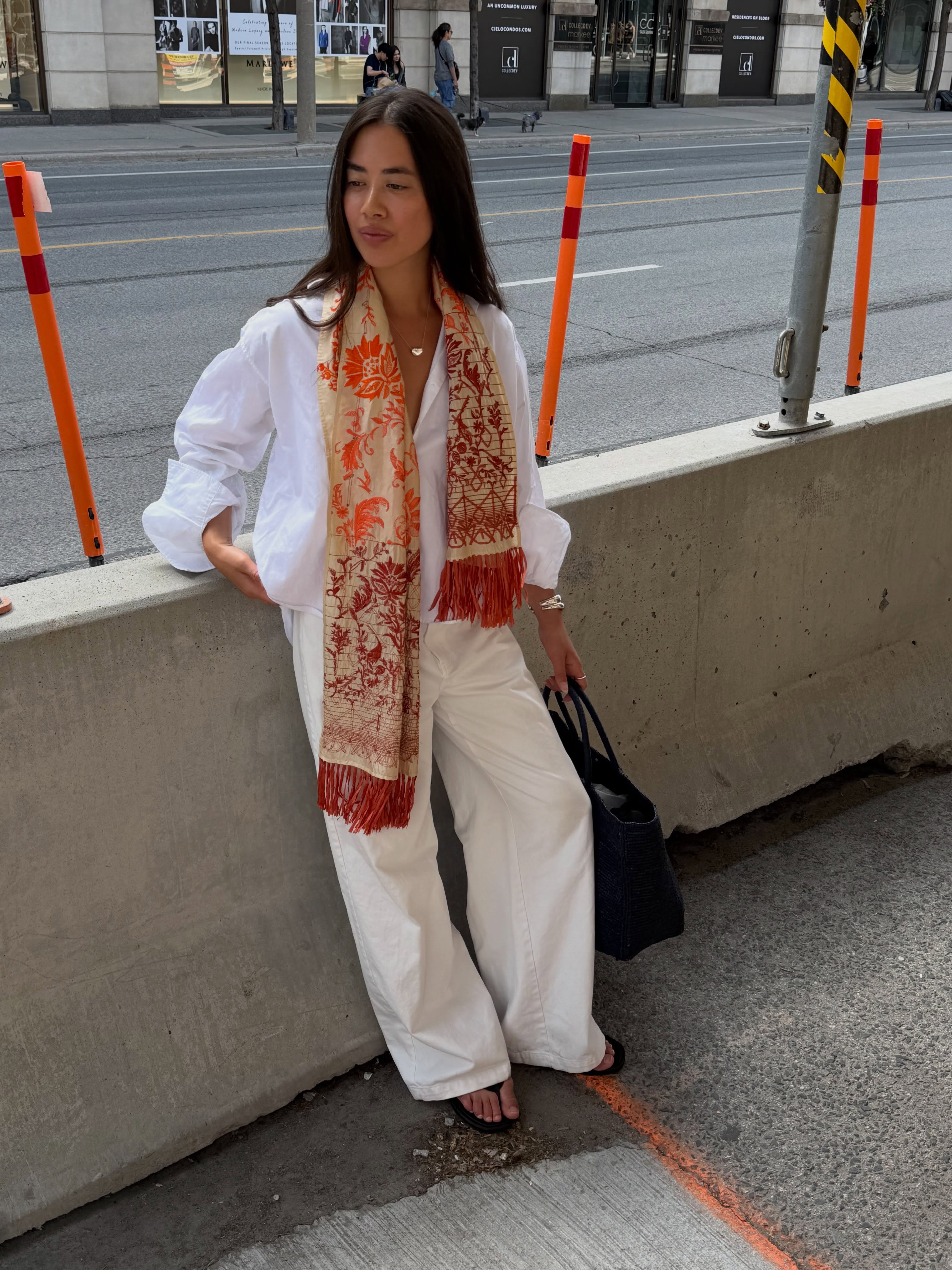 An image of an influencer leaning against a cement block wearing a white shirt, cream trousers and an orange, red and cream tassel scarf.