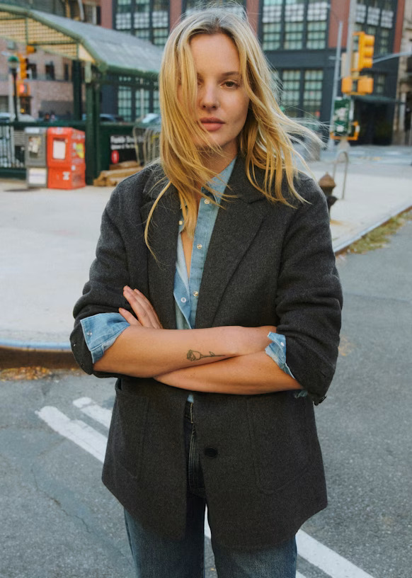 An image of a model wearing a grey wool blazer over a blue denim shirt standing in a street