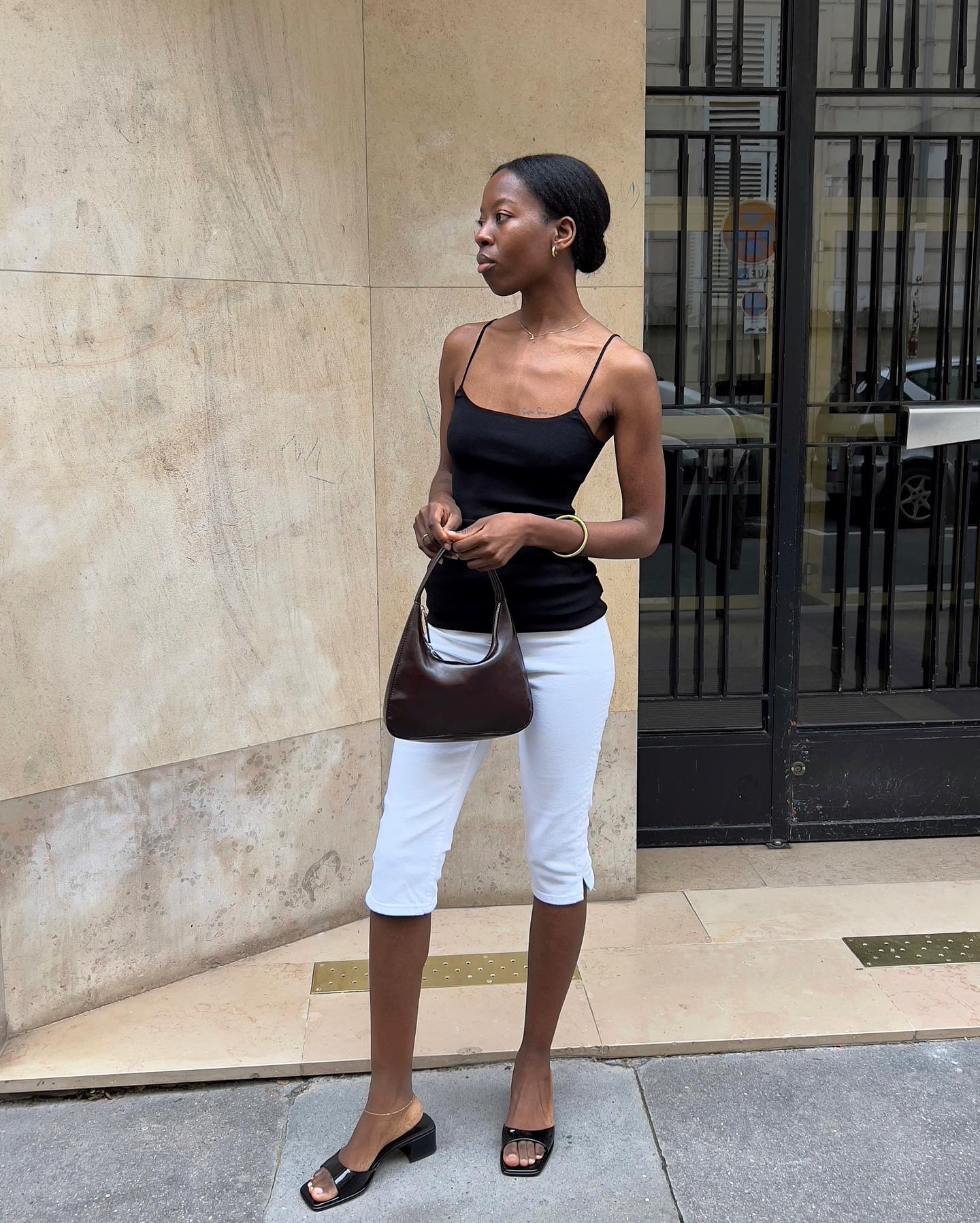An image of an influencer wearing a black camisole, white capri leggings and black mules holding a dark brown leather bag
