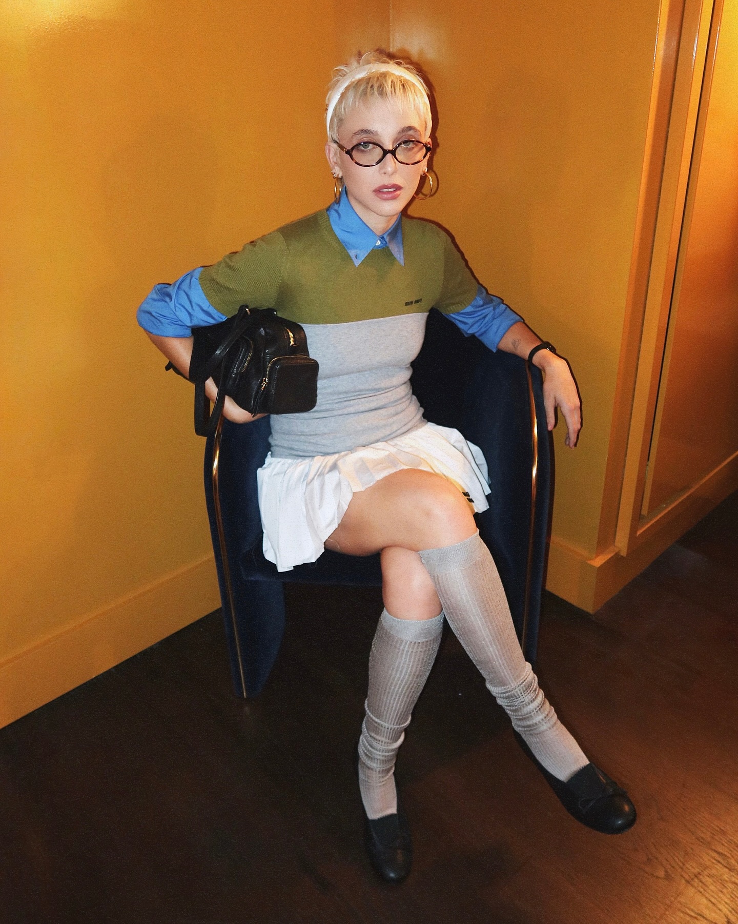 An image of influencer Emma Chamberlain wearing a Miu Miu outfit, with a white headband, brown glasses, a grey, green and blue long sleeve top, a white skirt, grey knee-high socks and black loafers