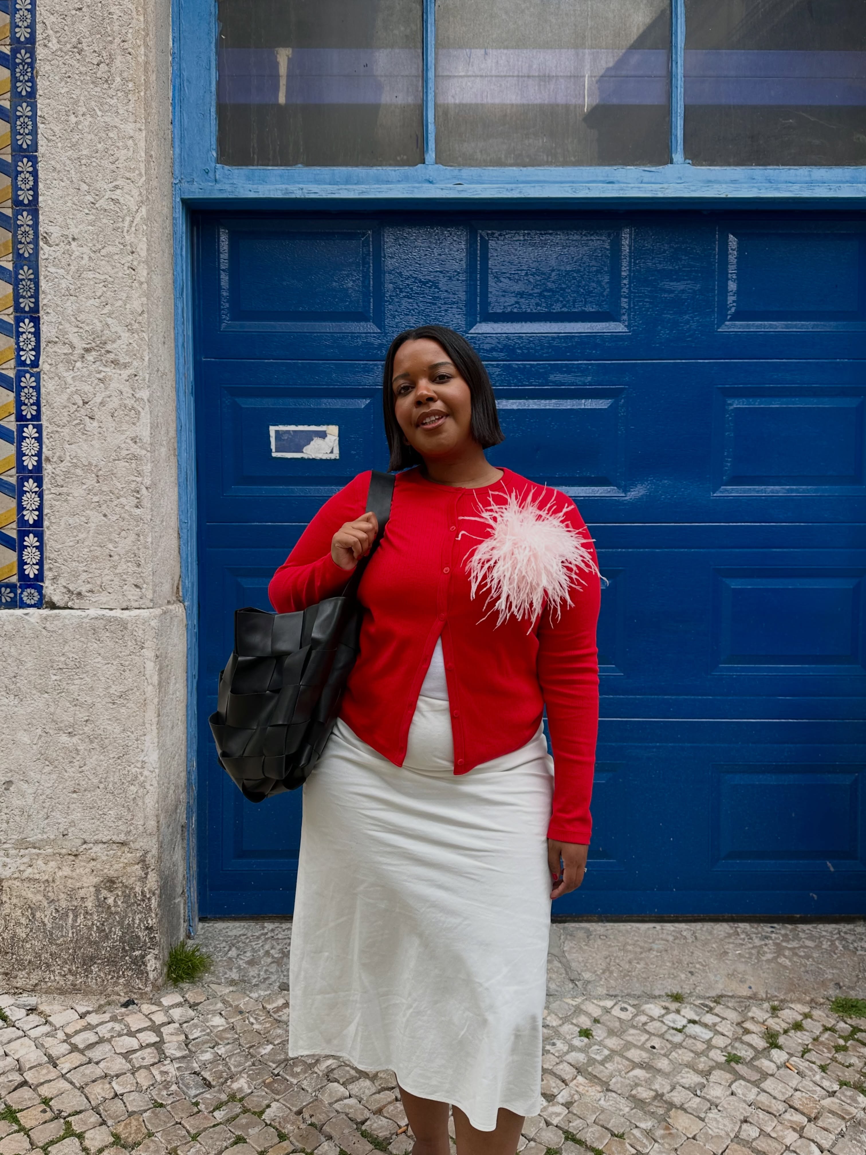 Influencer Karina wearing a red cardigan embellished with a pink feather brooch, with a white skirt and black bag.