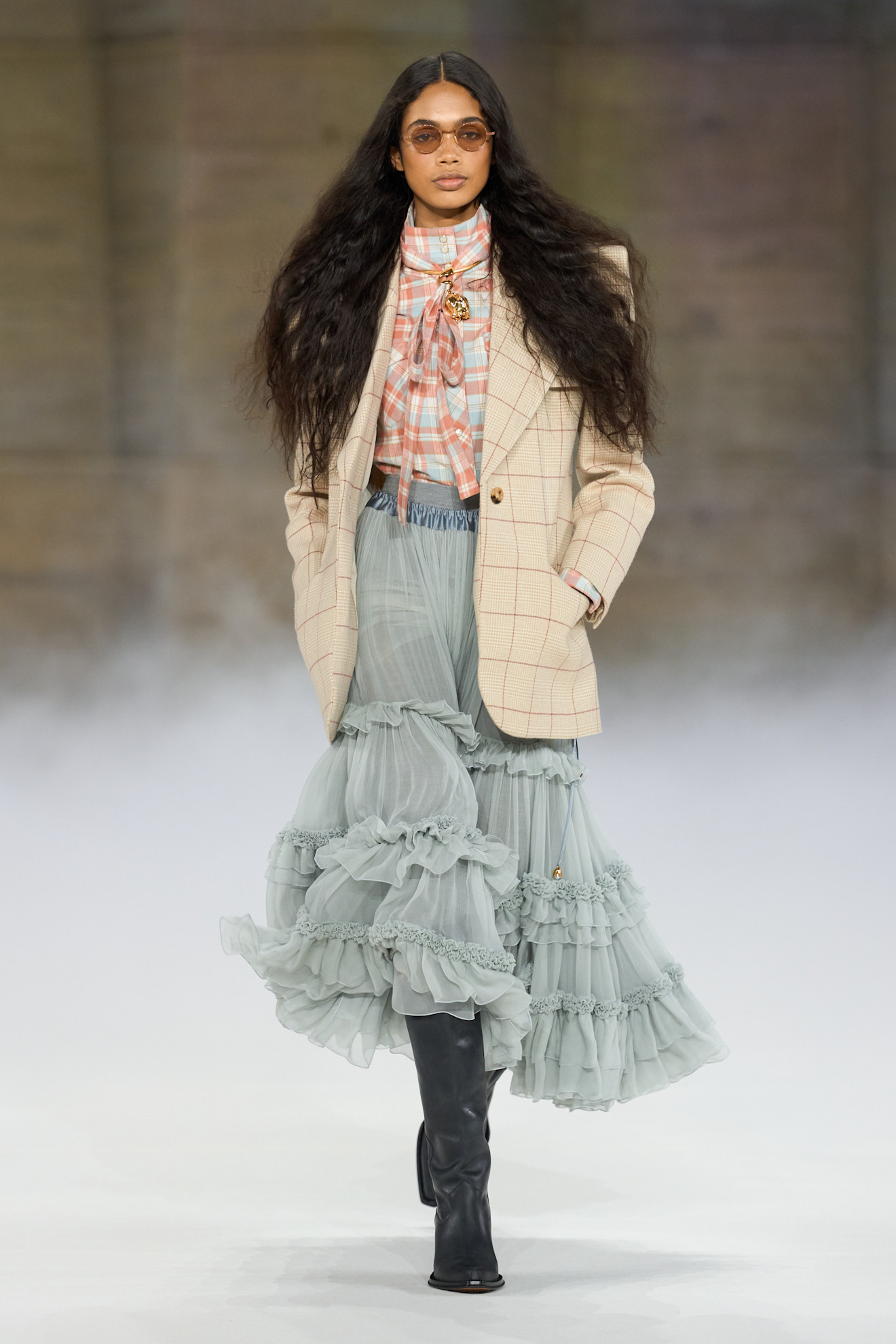 An image of a model from the autumn/winter 2026 fashion Chloé show wearing a plaid bow blouse, plaid beige blazer, dusty light blue ruffle skirt and black leather boots