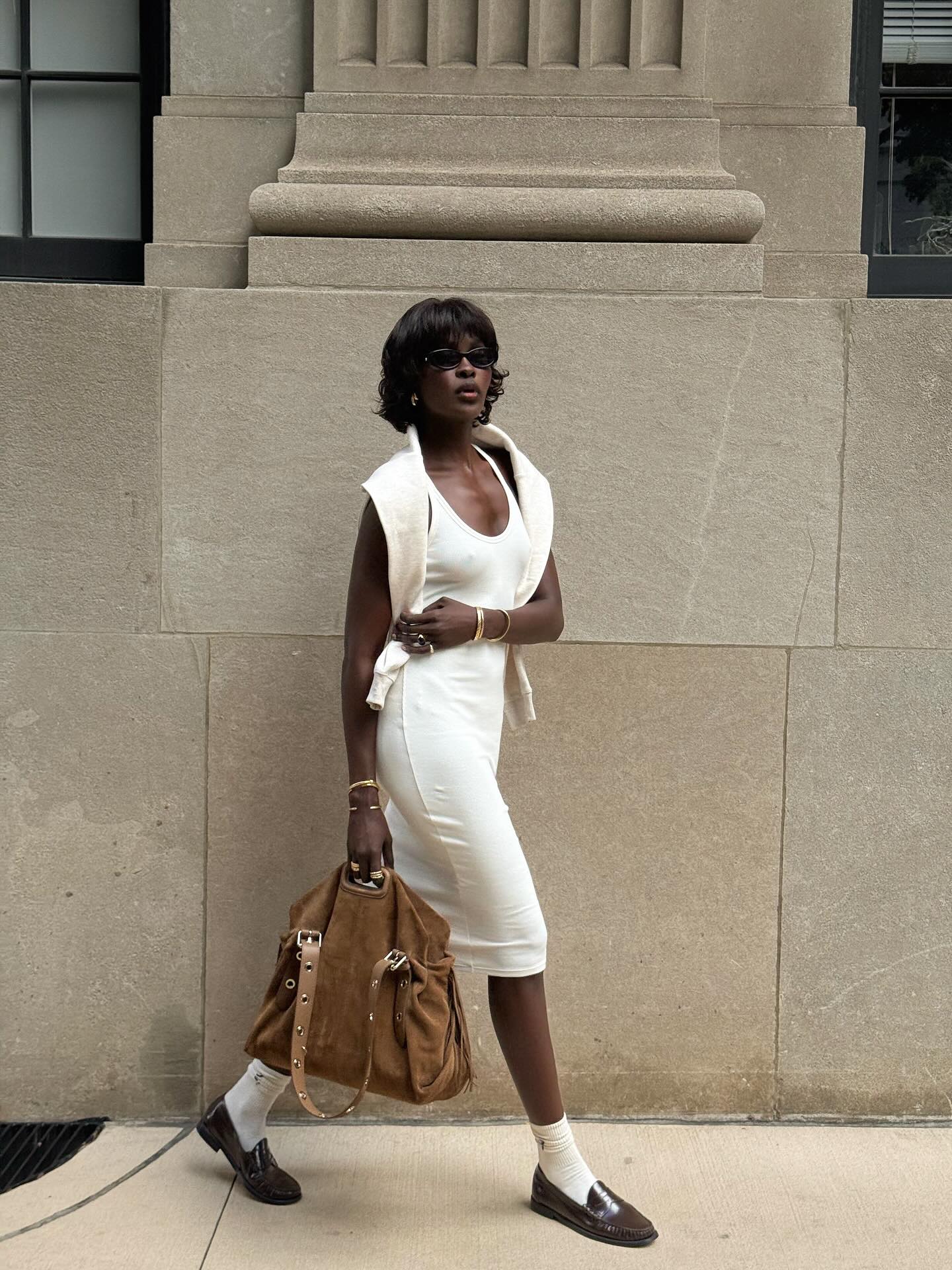 An image of an influencer wearing a white dress and brown loafers with stacked jewellery on her wrists and carrying a suede tote