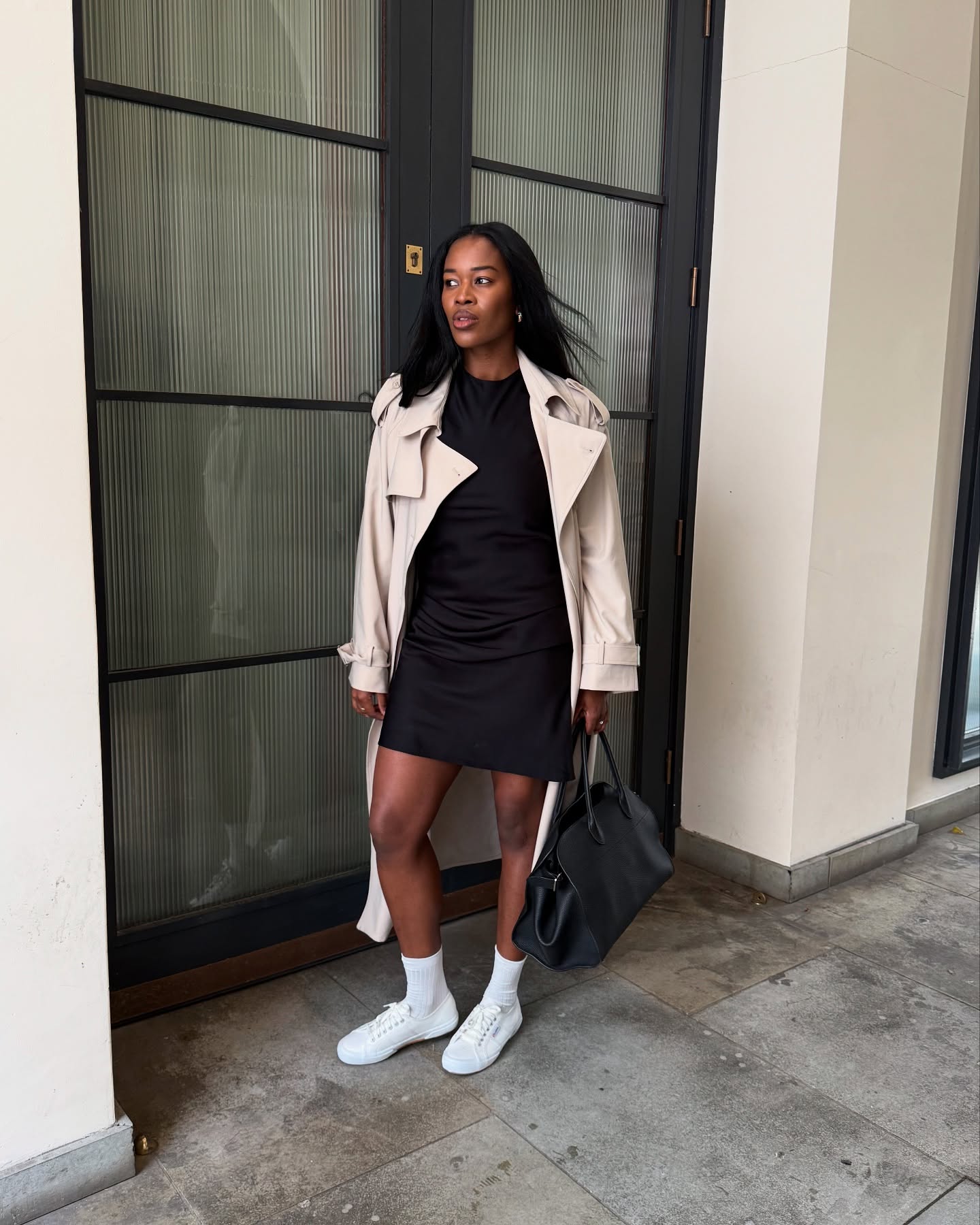 An image of an influencer wearing a black mini dress and a beige trench coat with white ankle socks and white trainers