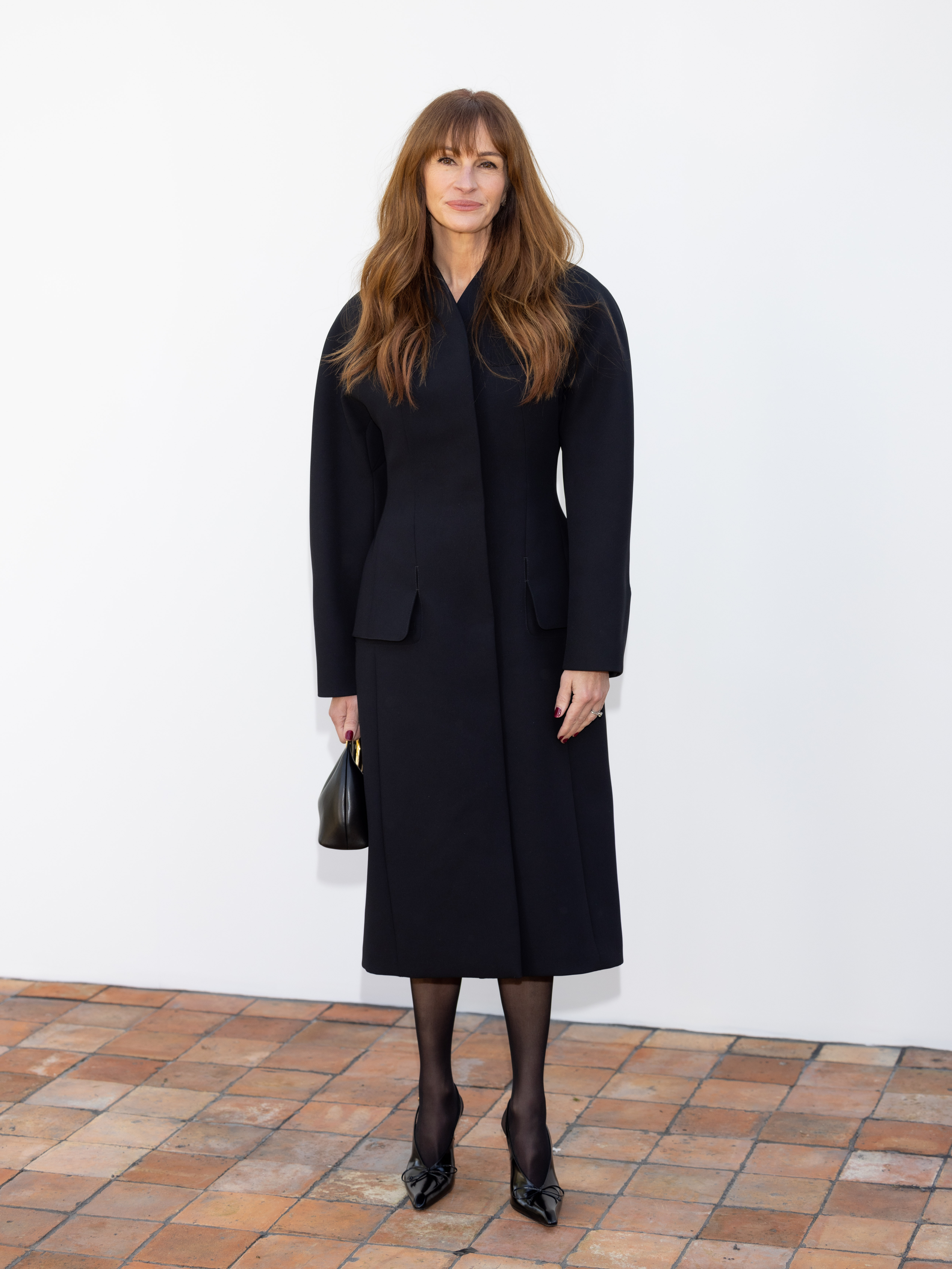 An image of actor Julia Roberts wearing a black coat with a nipped waist and black high heels, carrying a small black handbag