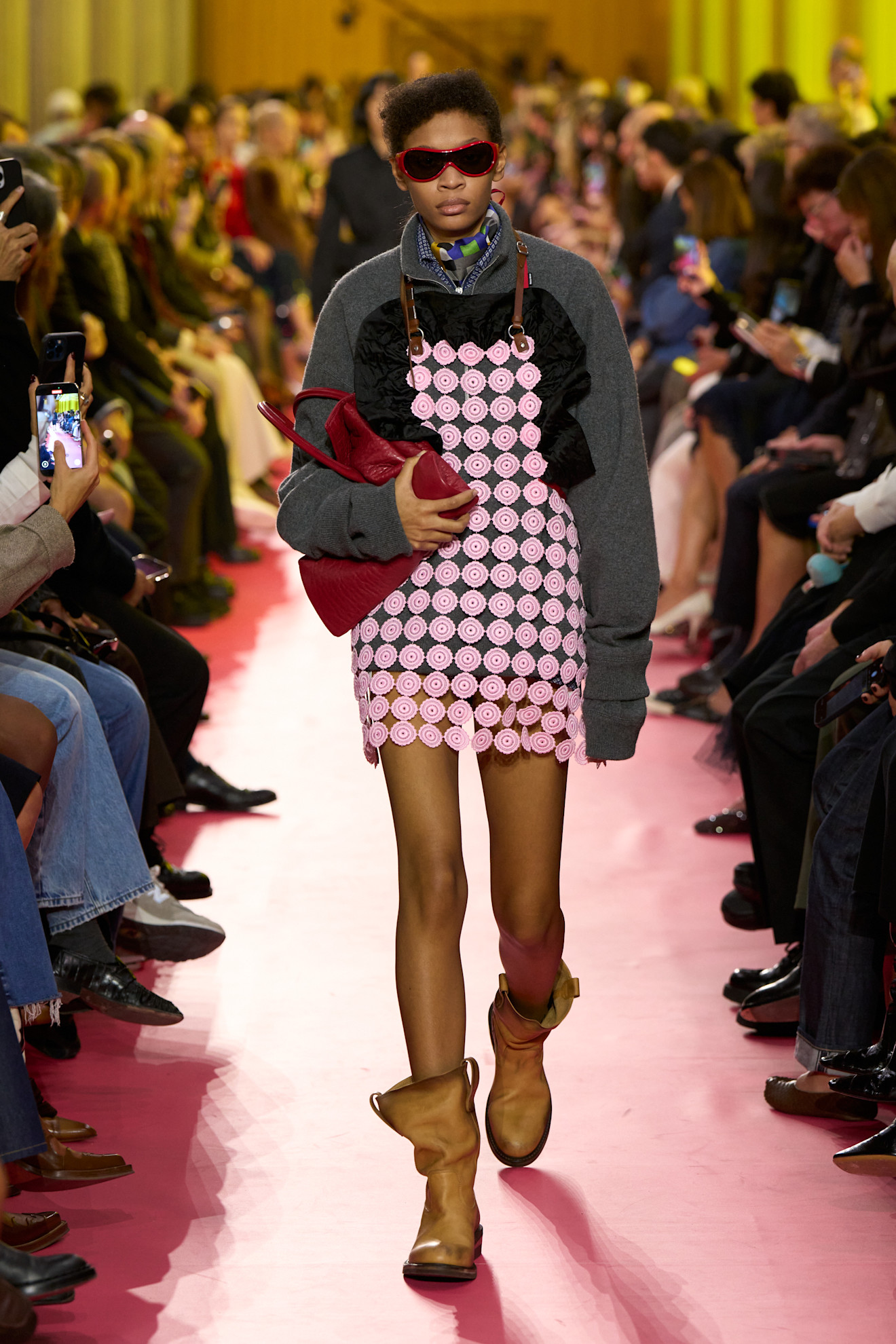 An image of a Miu Miu model wearing red sunglasses, a pink apron dress over a grey jumper and tan slouchy boots whilst carrying a red bag