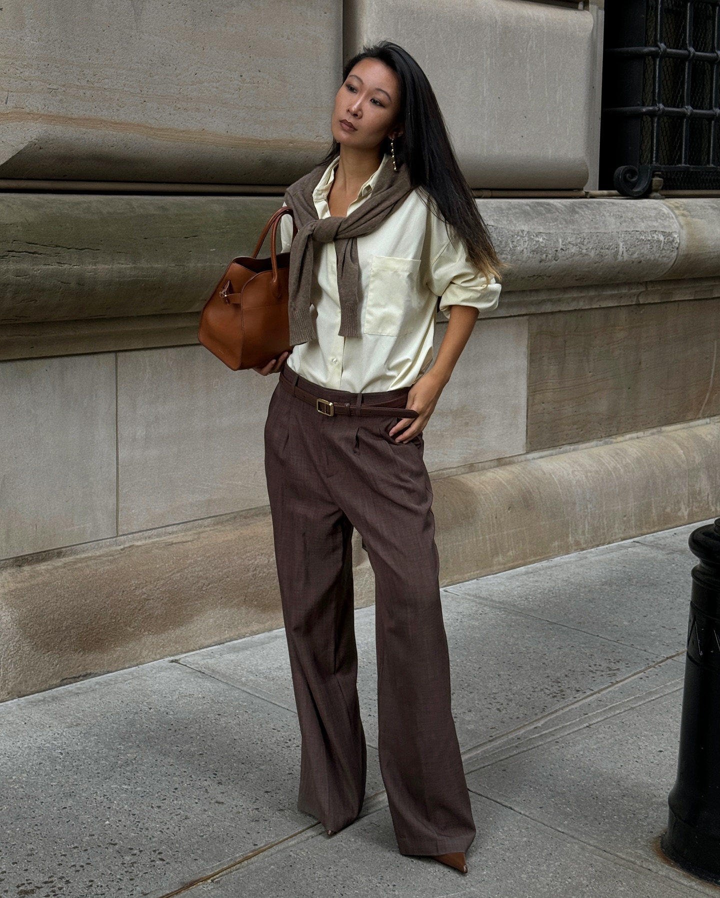 An image of an influencer wearing a pale yellow button-down, grey trousers, a sweater wrapped around her shoulders and holding a brown tote in New York City
