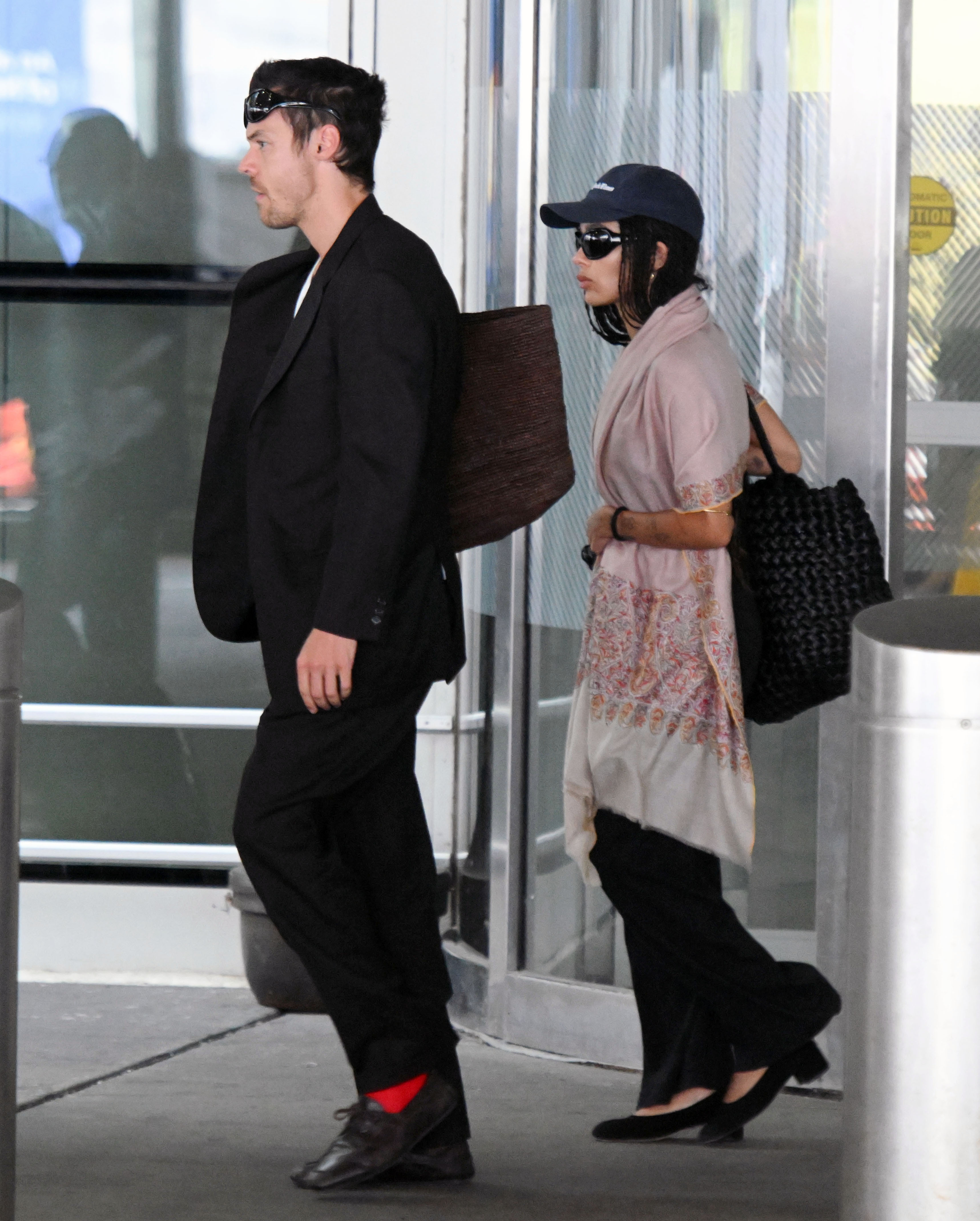 Zoe Kravitz and Harry Styles emerge from the airport. Zoe wears black trousers with heeled ballet pumps and a pink throw over her shoulders.