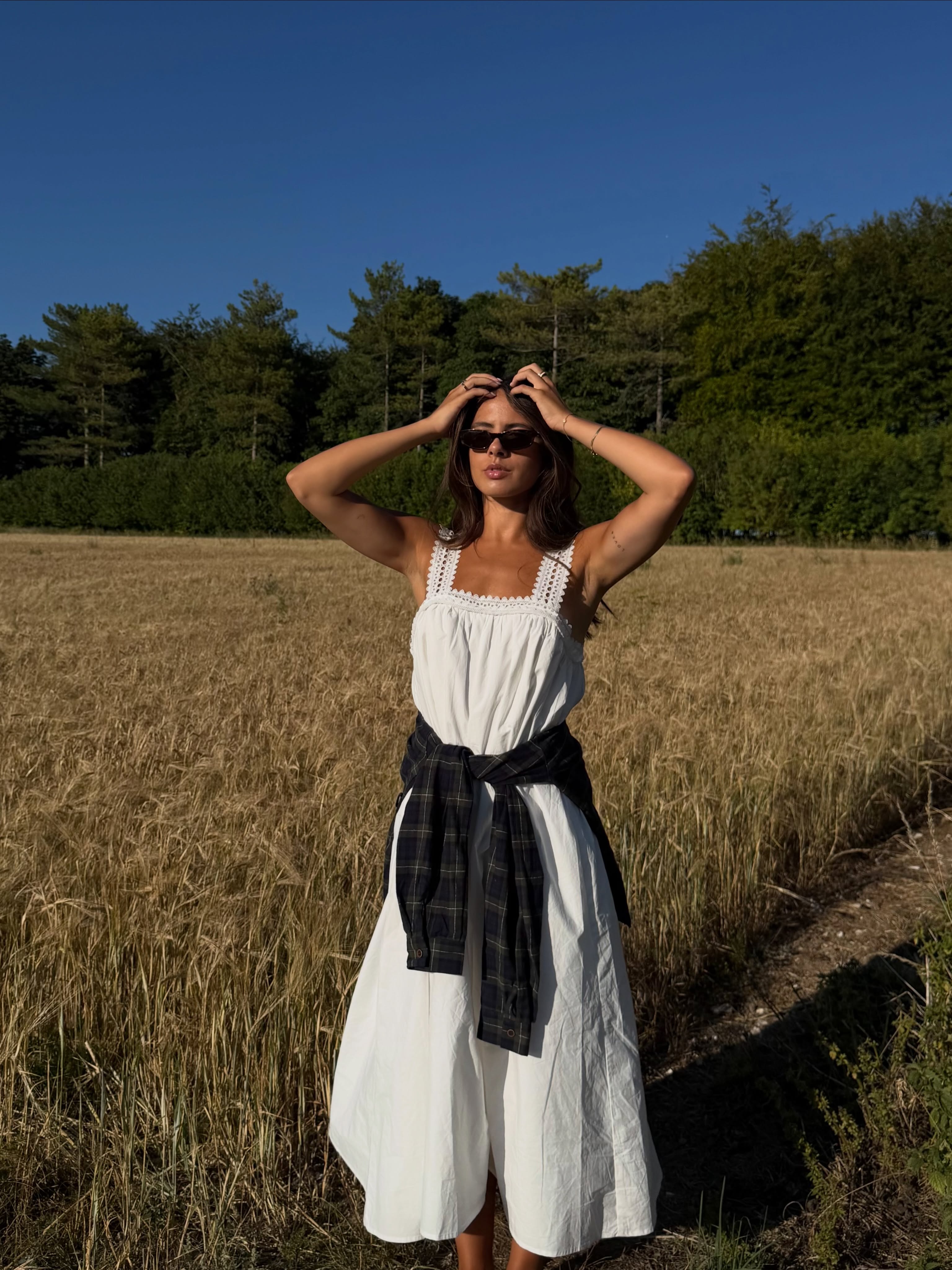 Influencer Tia Dewitt wearing a white broderie anglaise dress with a plaid shirt and sunglasses.